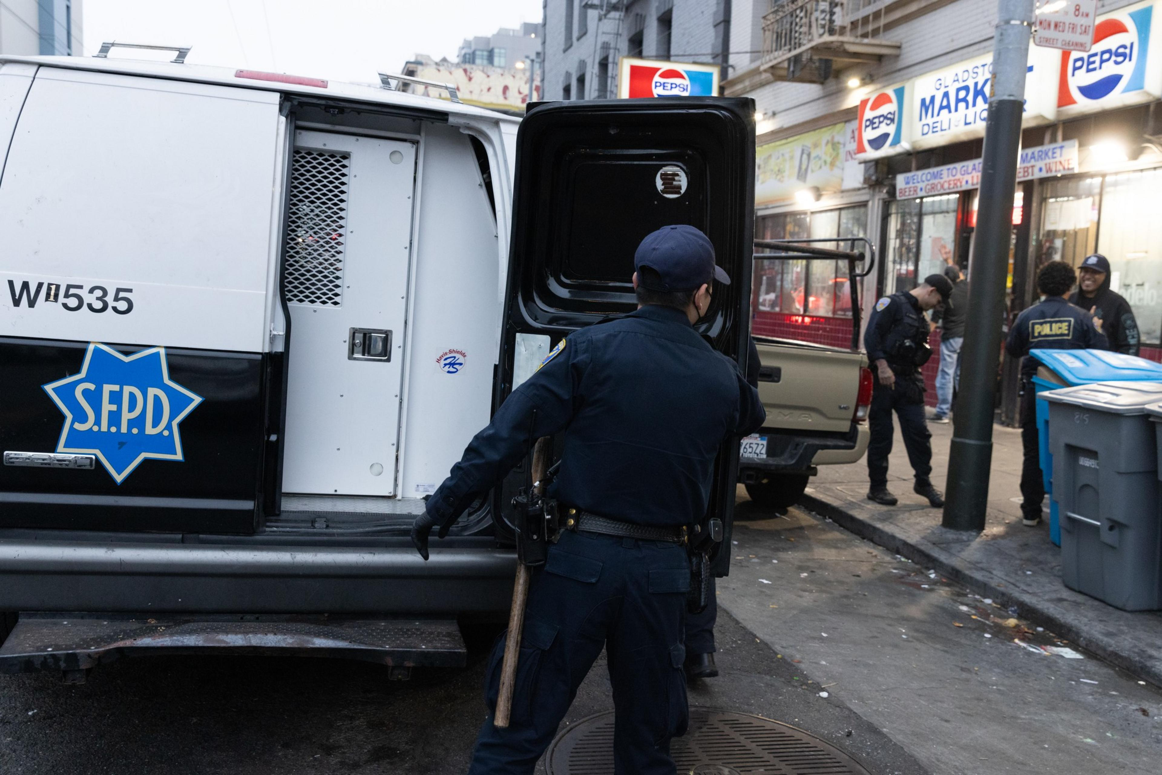 A police officer stands by an open SFPD van on a city street. Nearby, more officers are conversing with people outside a market under Pepsi signs.