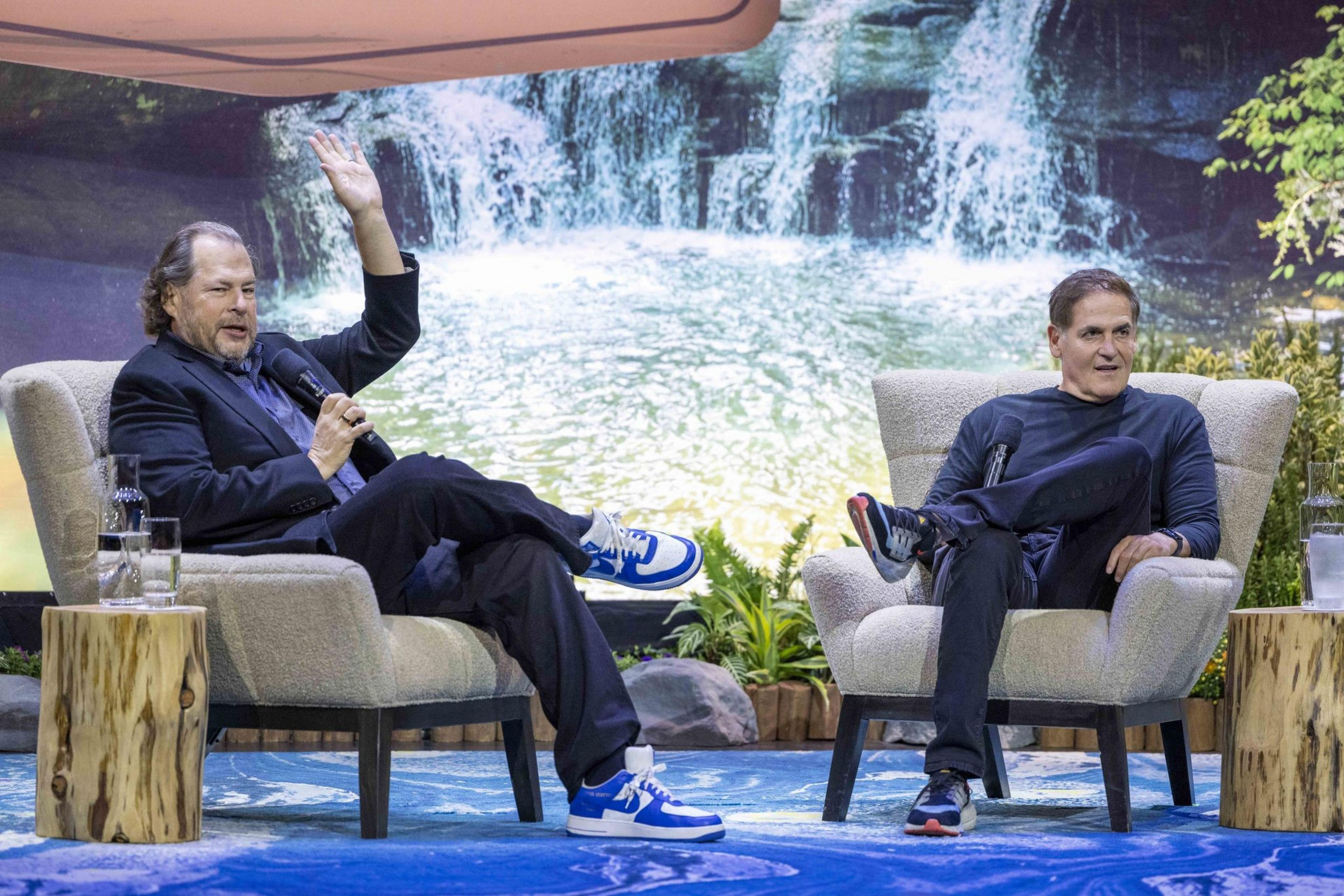 Two men sit in armchairs on a stage with a waterfall backdrop. One, in a suit and blue sneakers, raises his hand while another, in casual attire, looks at him.