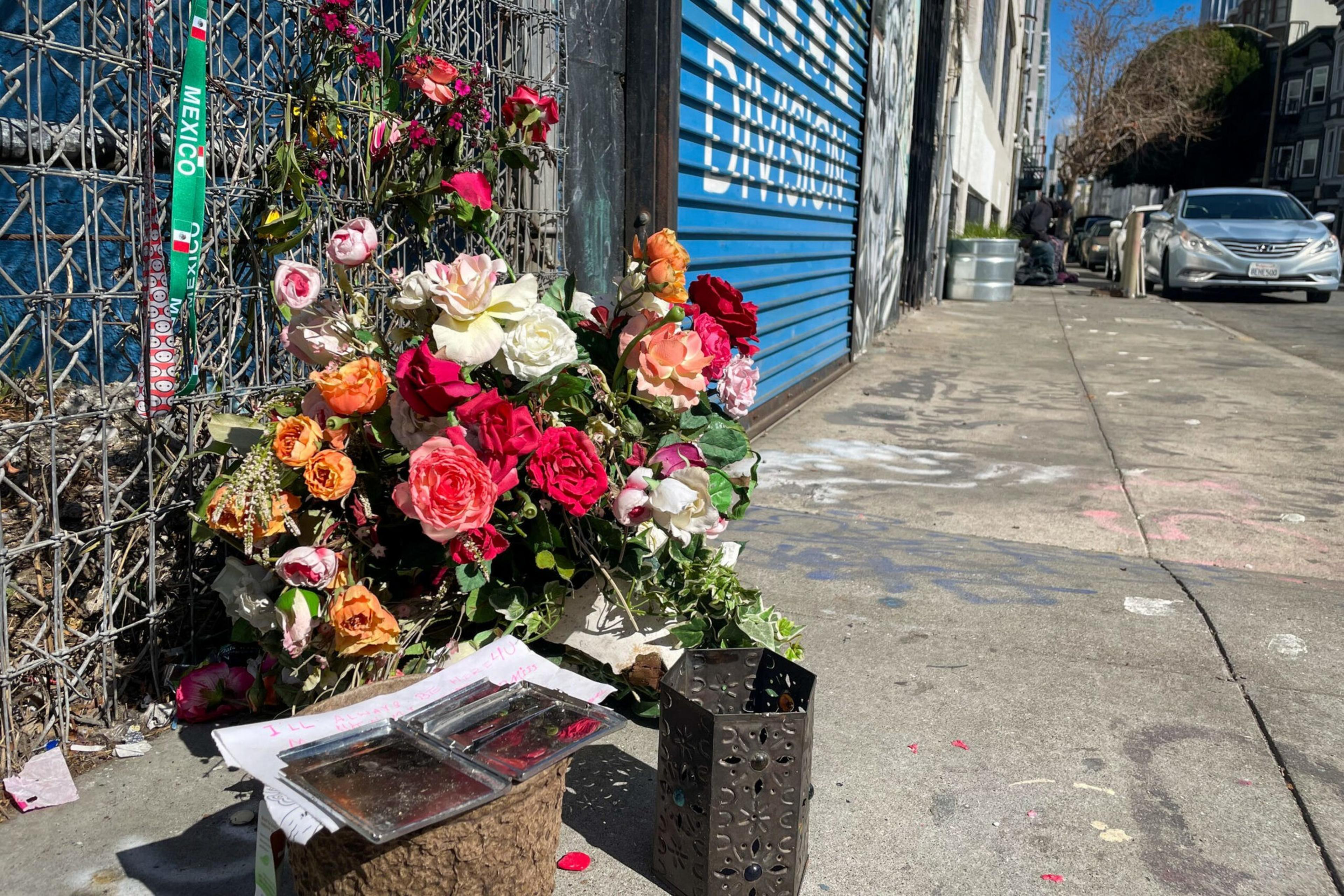 A colorful bouquet of flowers rests against a fence on a sunlit sidewalk, next to a metal container and some scattered papers near a blue garage door.