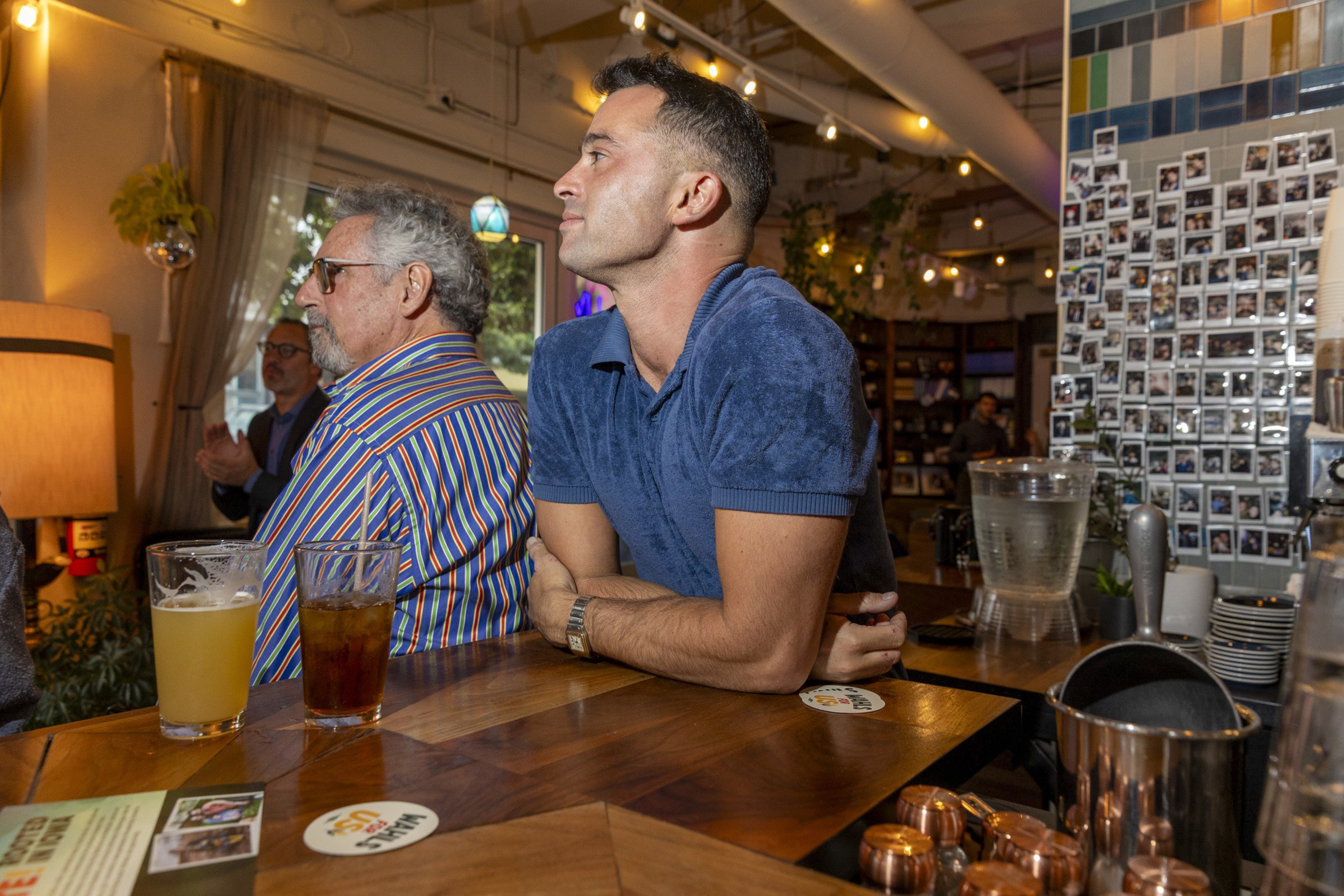 Two men sit at a wooden bar, one in a striped shirt and the other in a blue polo, with drinks in front of them and a wall covered in photos behind.