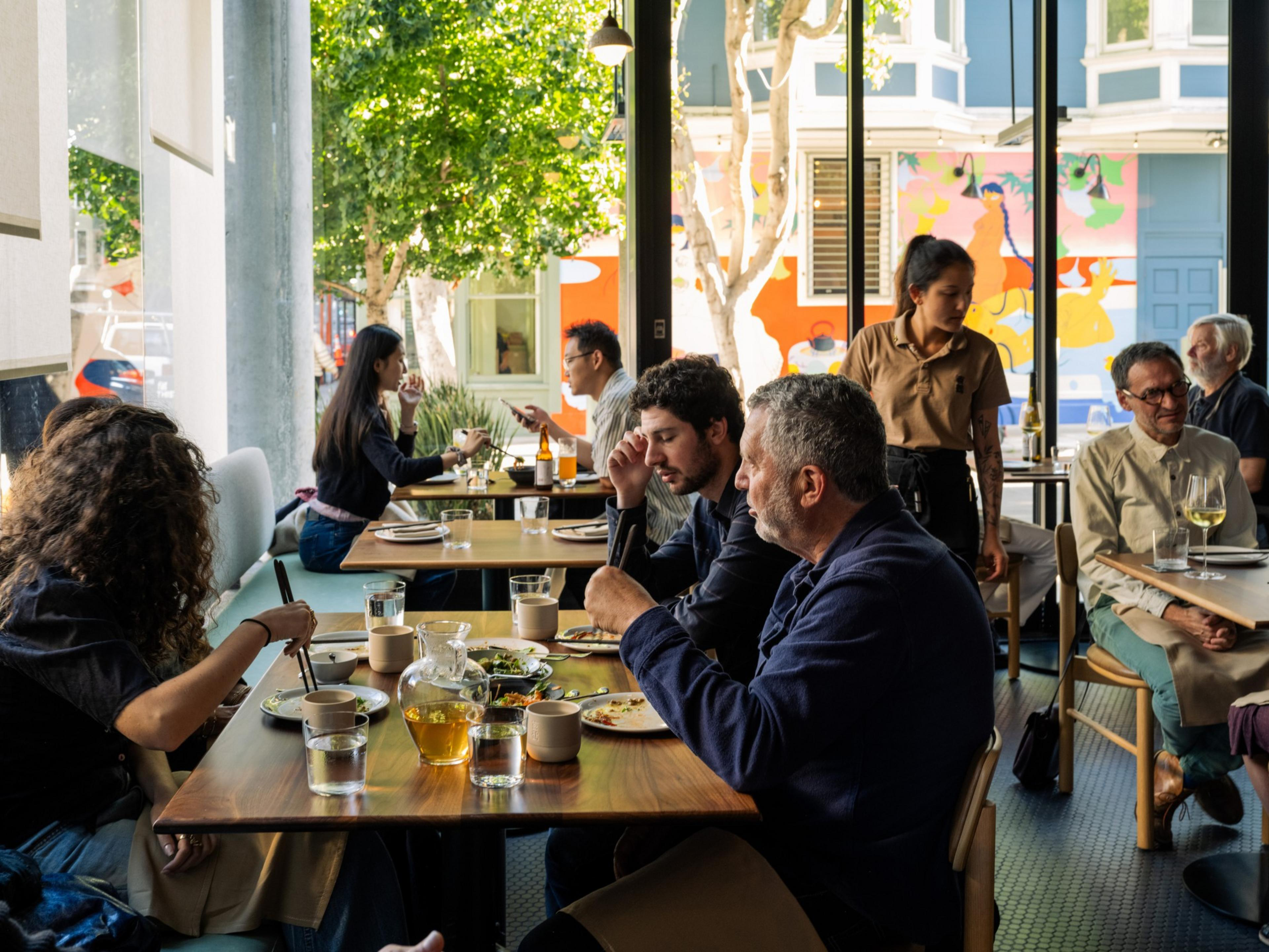 People are dining at a modern restaurant with large windows, enjoying food and drinks while a server attends to a table.