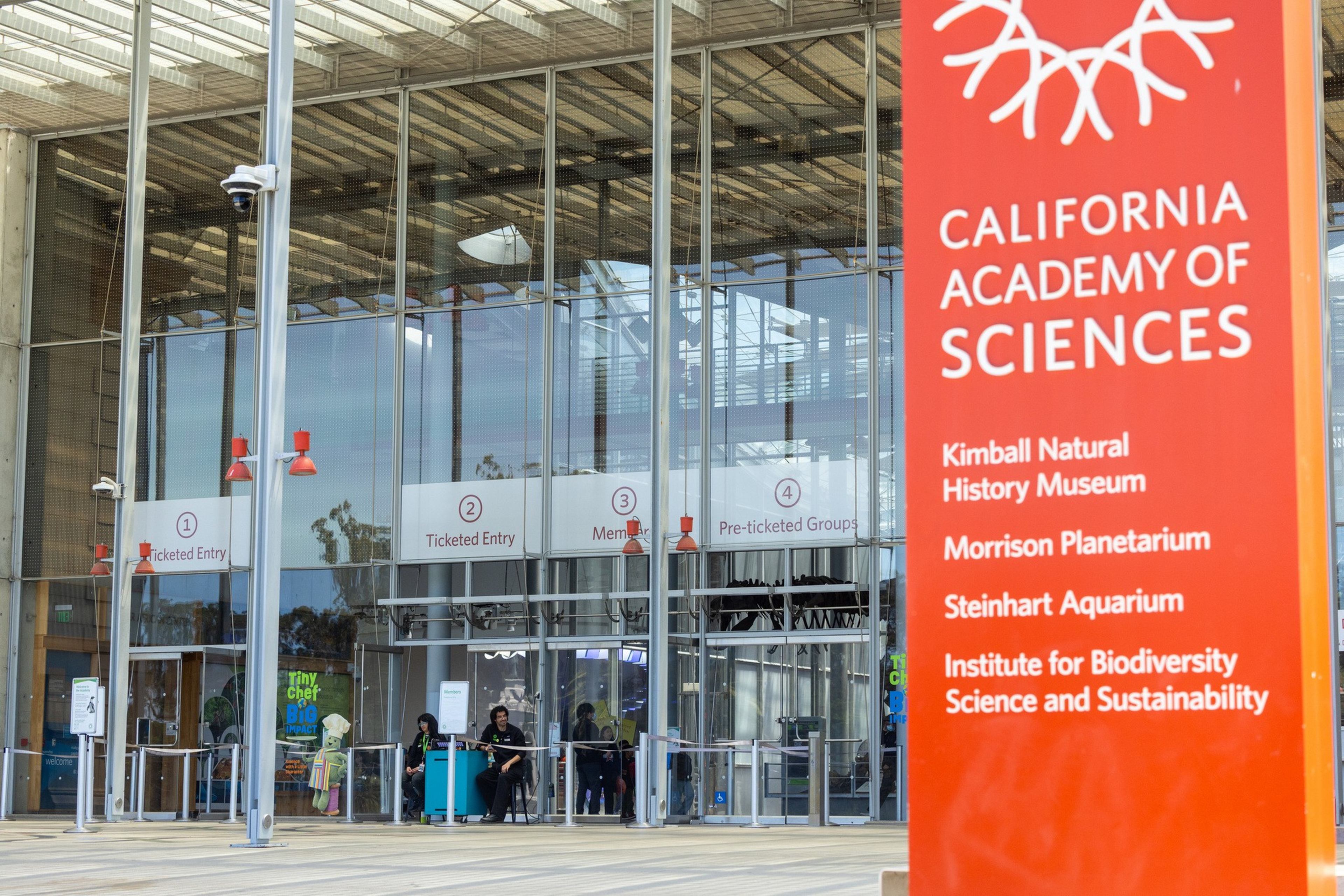 A large red sign lists California Academy of Sciences and its exhibitions beside glass entrances labeled for ticketed entry and members.