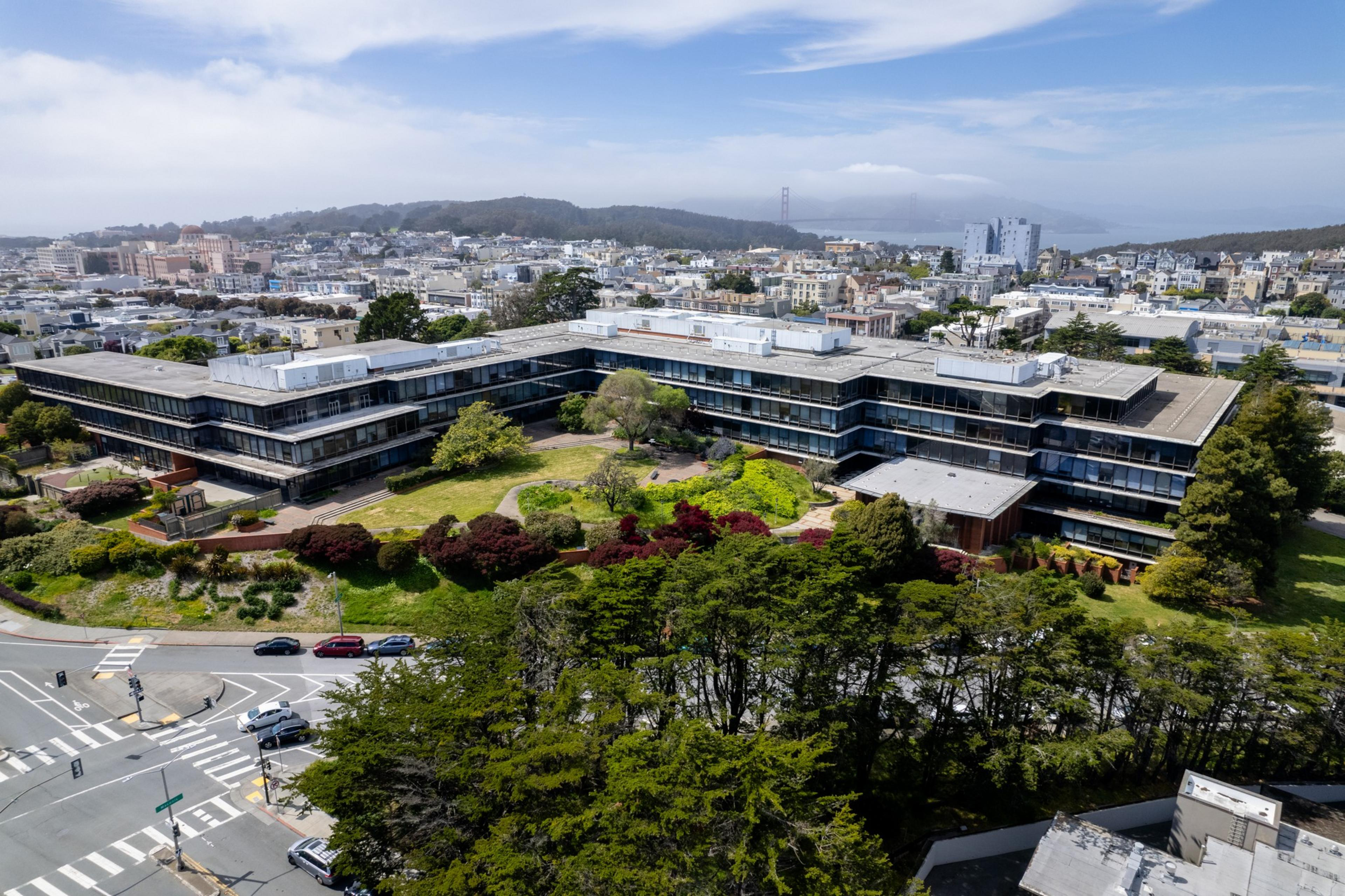 Aerial view of a modern building with terraces overlooking a cityscape with trees and distant hills.