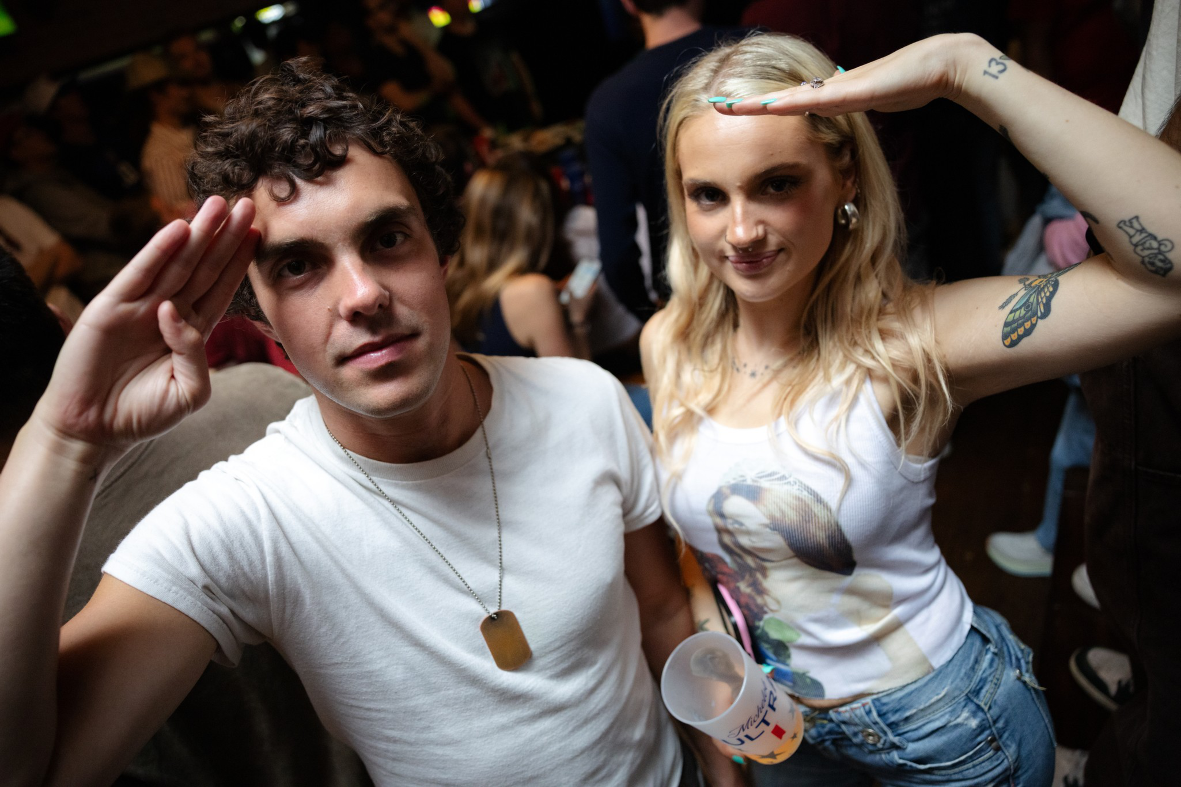 A young man and woman pose with salutes; the man wears a white shirt and dog tag, the woman has blonde hair, tattoos, a tank top, and holds a cup.
