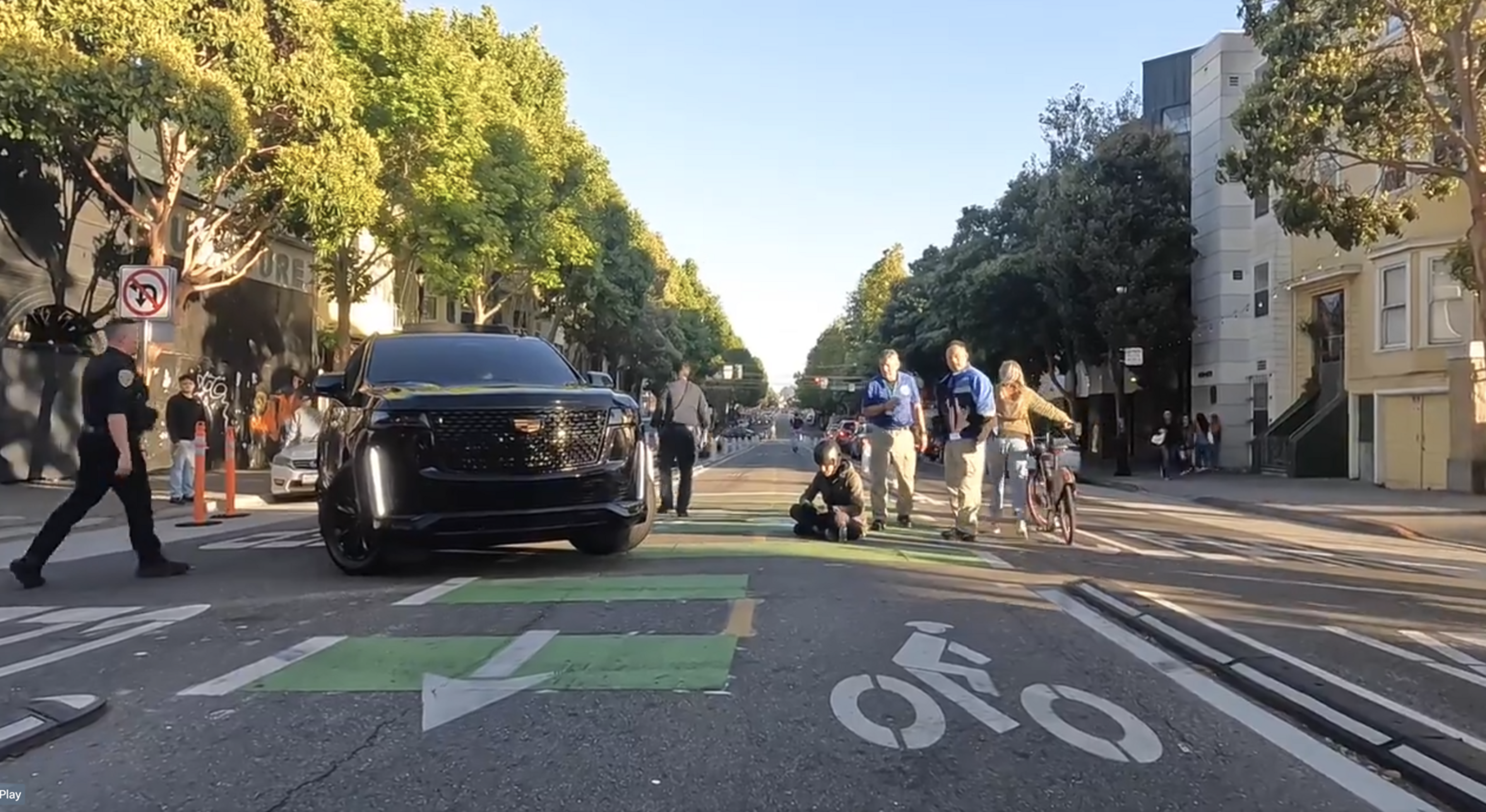 A black SUV is stopped in the middle of a city street while pedestrians and a cyclist cross. A police officer walks nearby, and the street is lined with trees and buildings.
