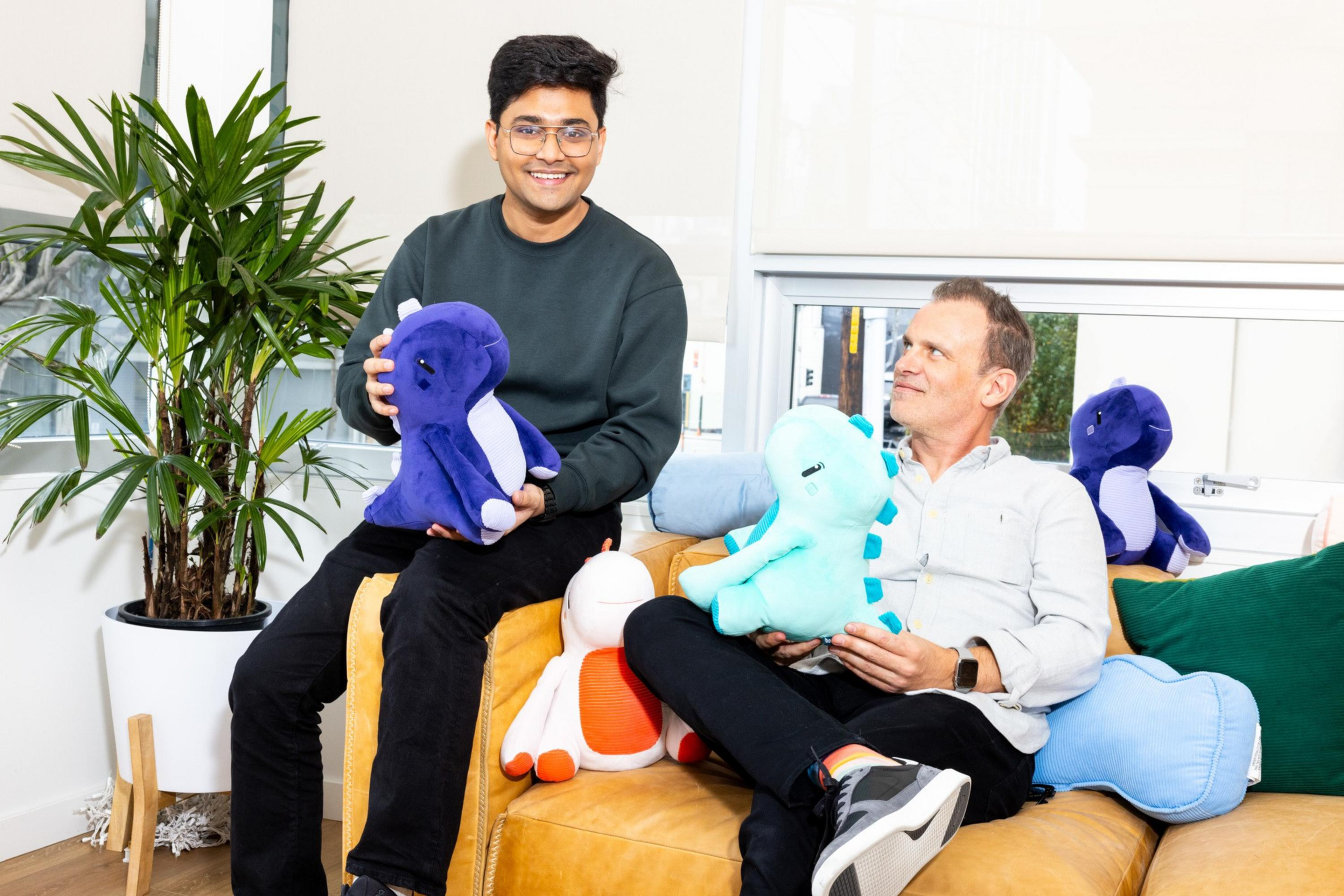 Two men sit on a yellow couch, each holding a colorful dinosaur plush toy, surrounded by more plush toys, with a large potted plant and bright window behind them.