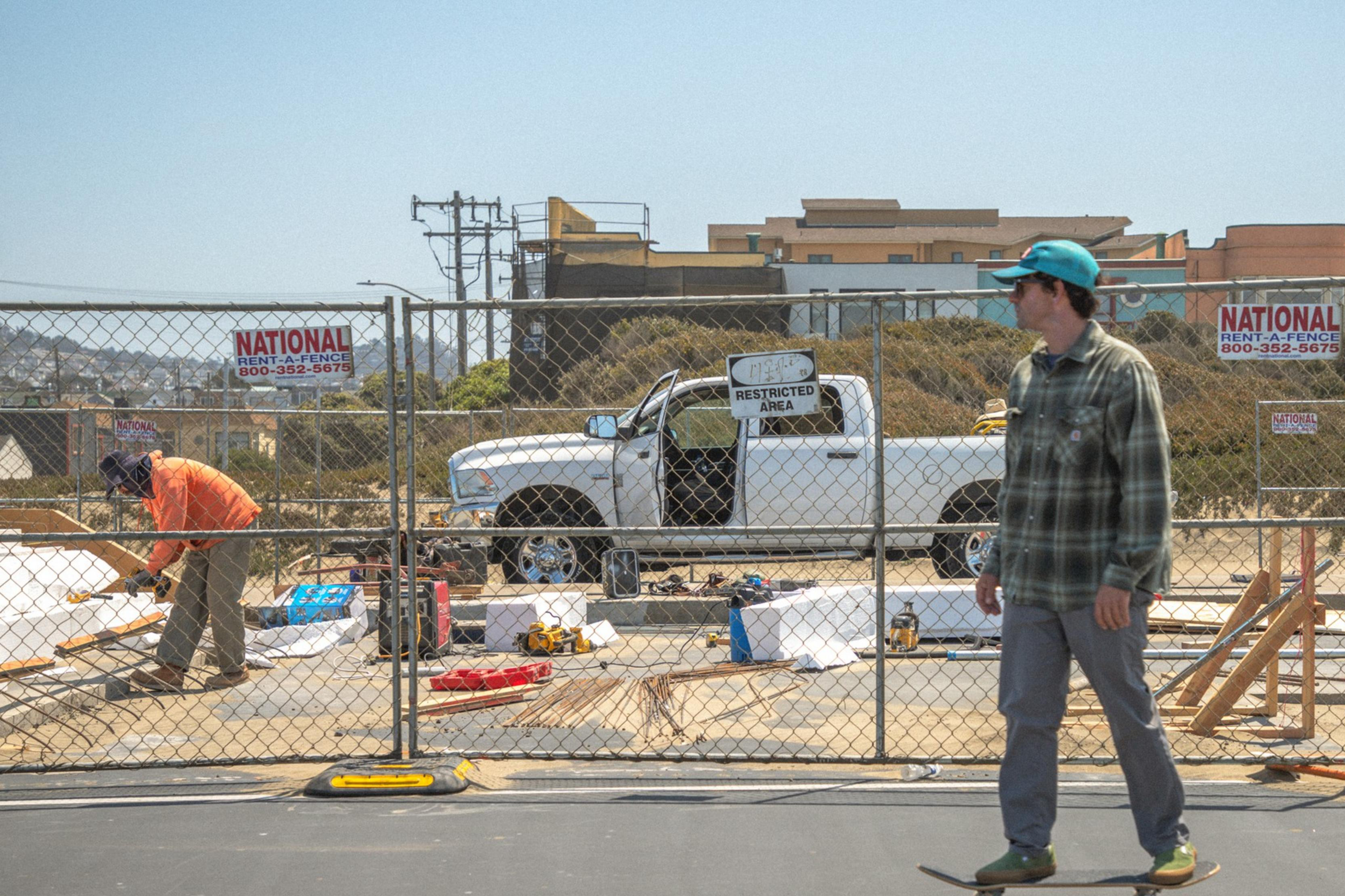 A man skateboards past a construction site, where another person is working near a pickup truck. A fence with "Restricted Area" signs surrounds the site.
