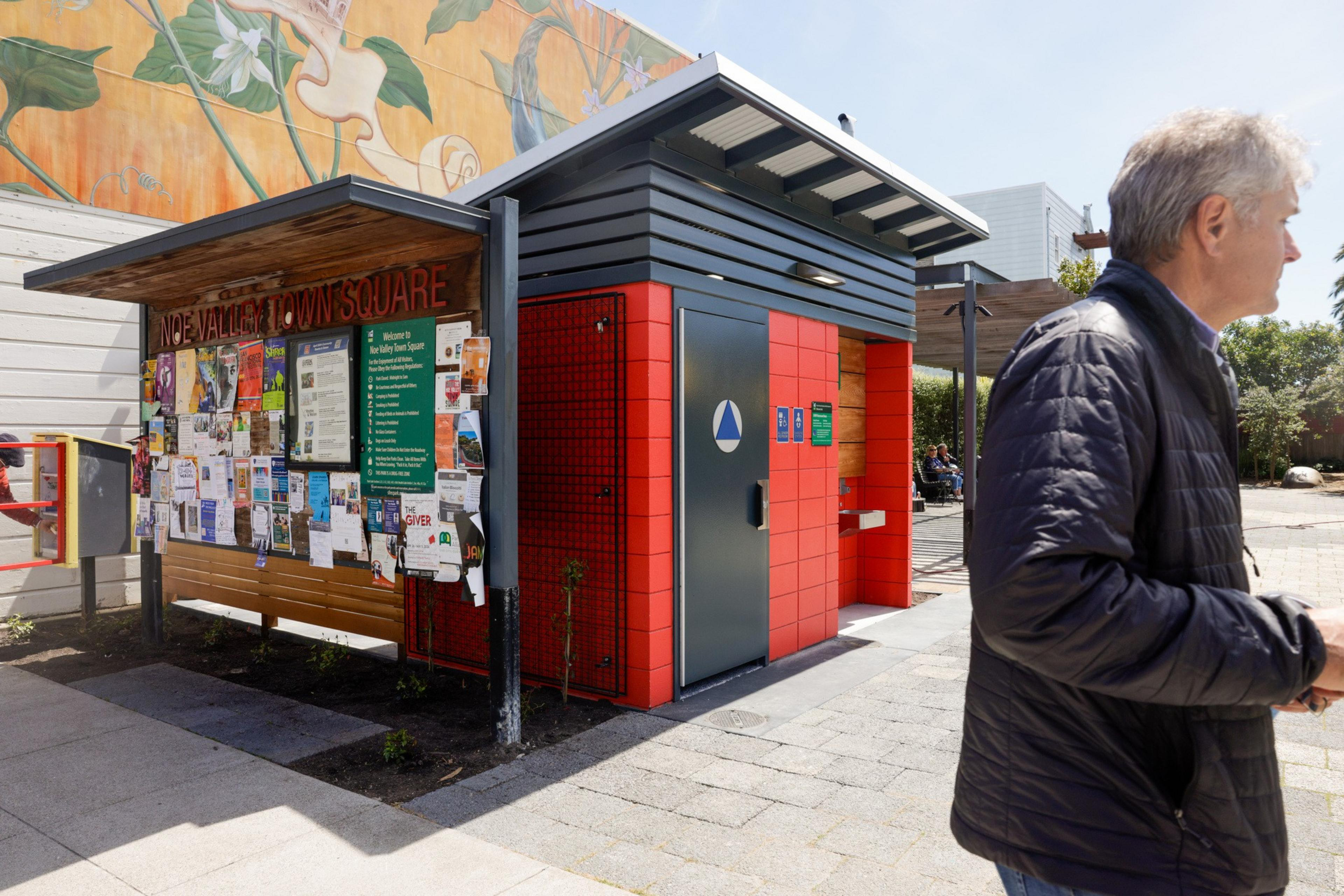 A modern public restroom with red accents by a community bulletin board, under a flowering tree mural. A man walks by in the foreground.