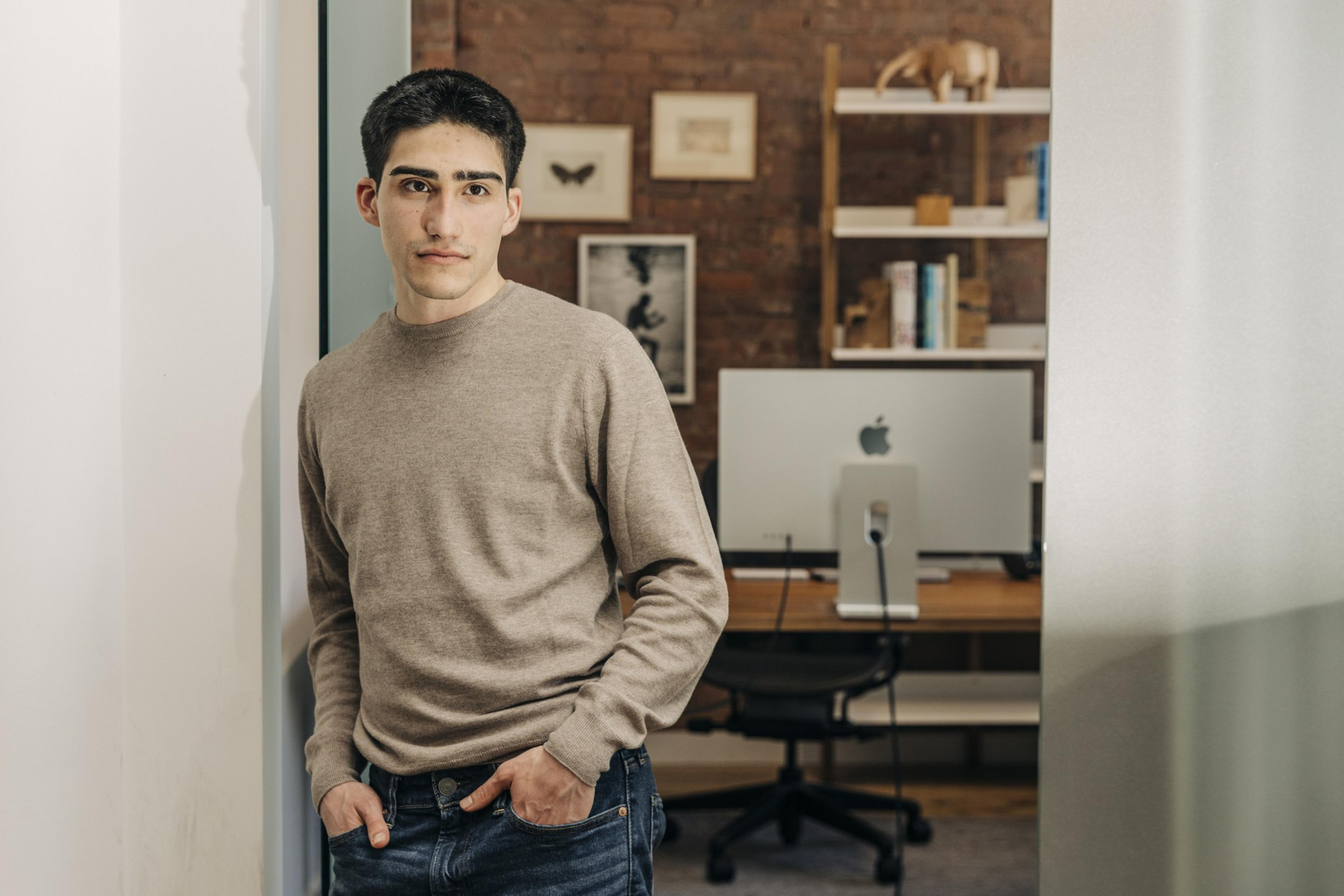 A young man in a brown sweater and jeans stands with hands in pockets in a modern office with a computer and a shelf in the background.