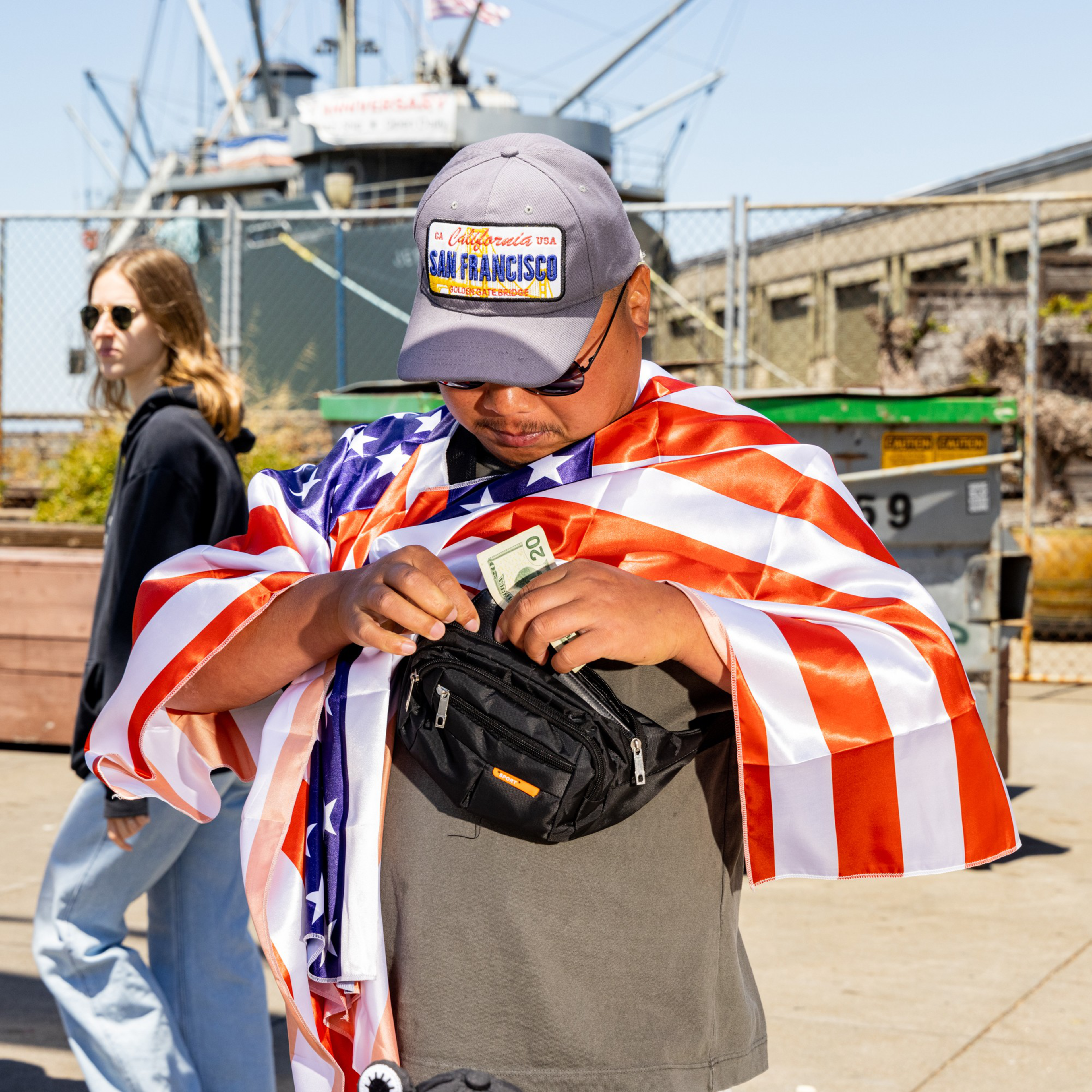 A man, draped in an American flag, wearing a San Francisco cap, retrieves a $20 bill from a black waist pouch. There's a ship and another person in the background.