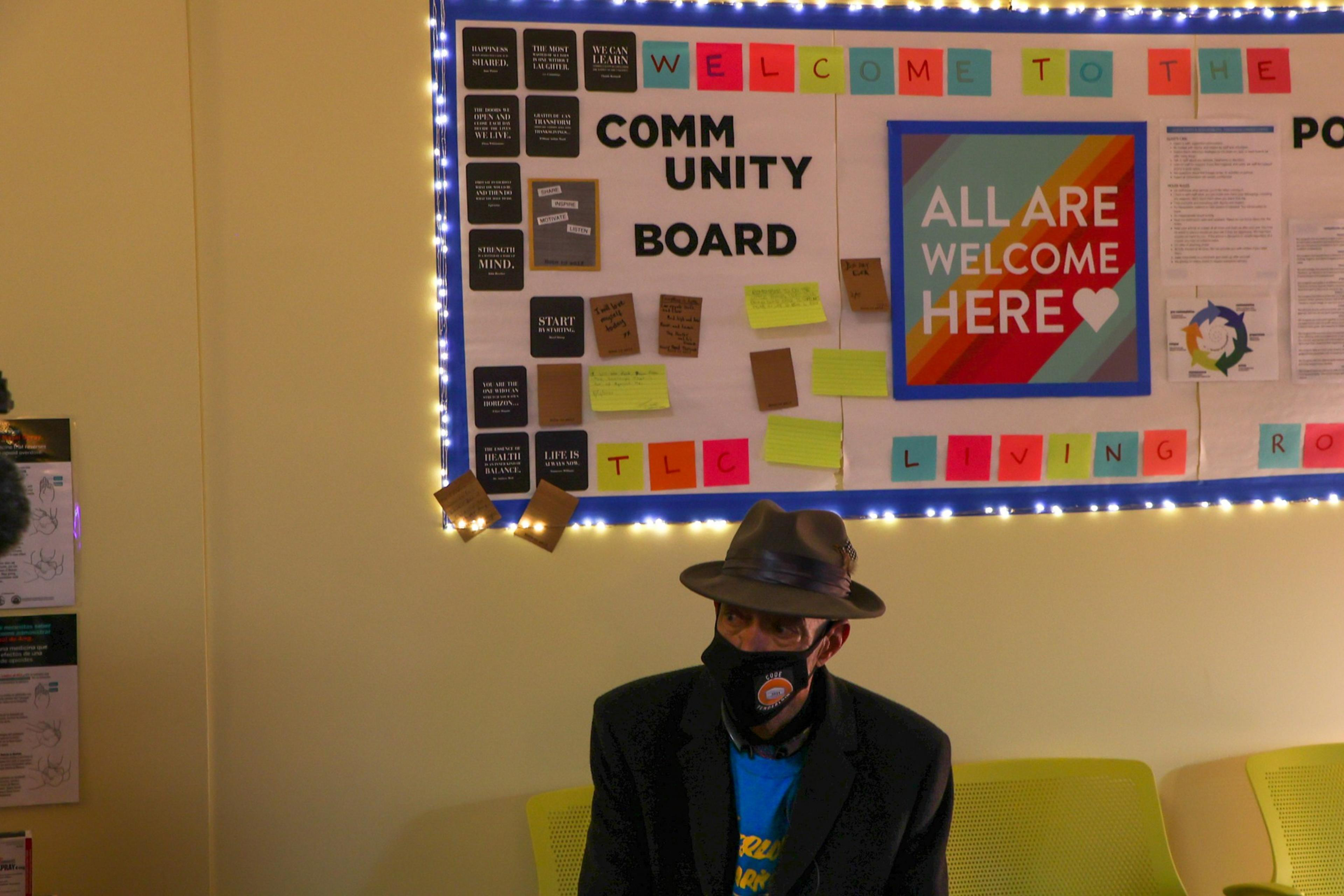 A man wearing a fedora and face mask sits on a yellow bench beneath a brightly lit community board with colorful notes and a sign reading "All Are Welcome Here."