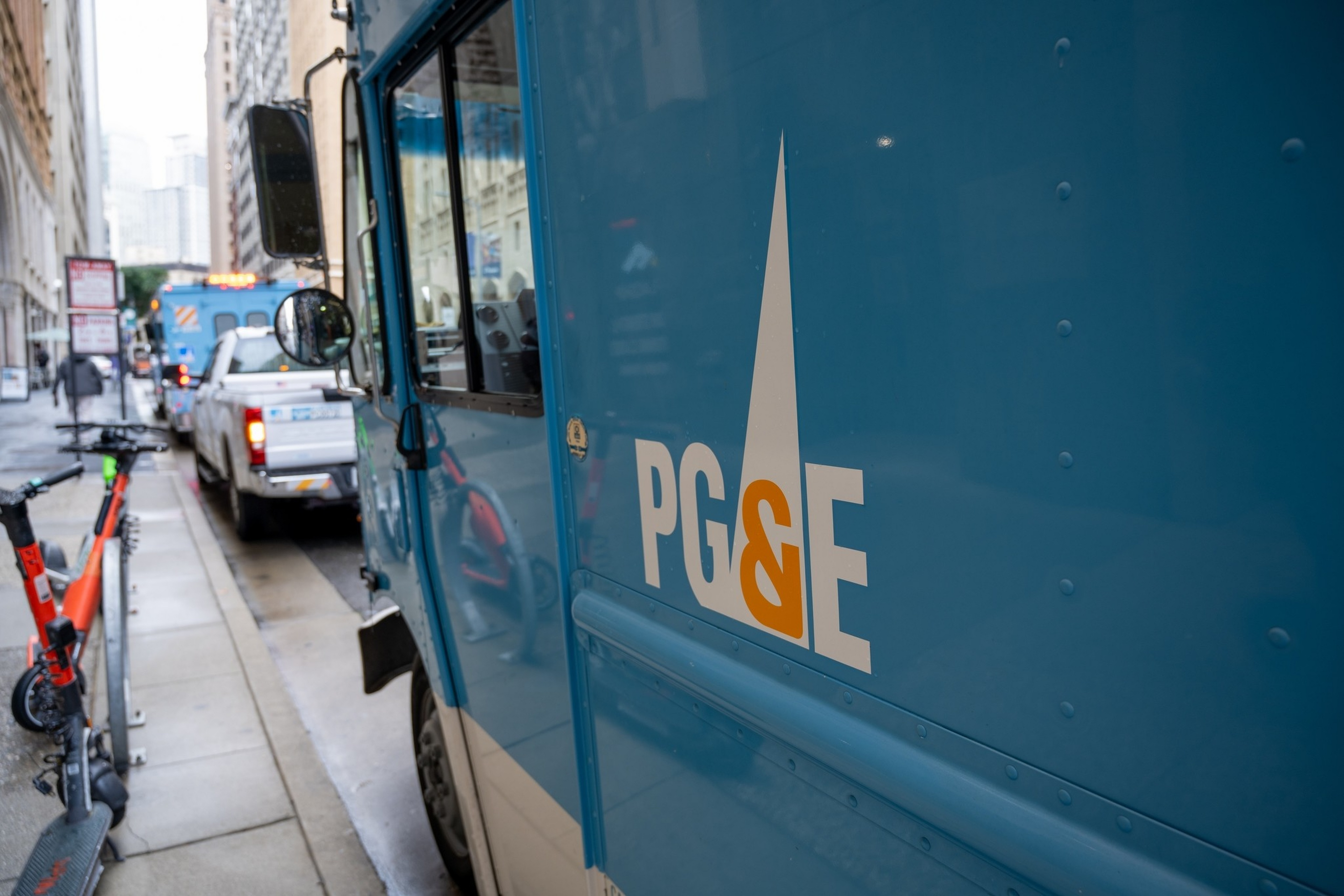 A blue PG&E utility truck is parked on a city street with electric scooters lined up on the sidewalk nearby.