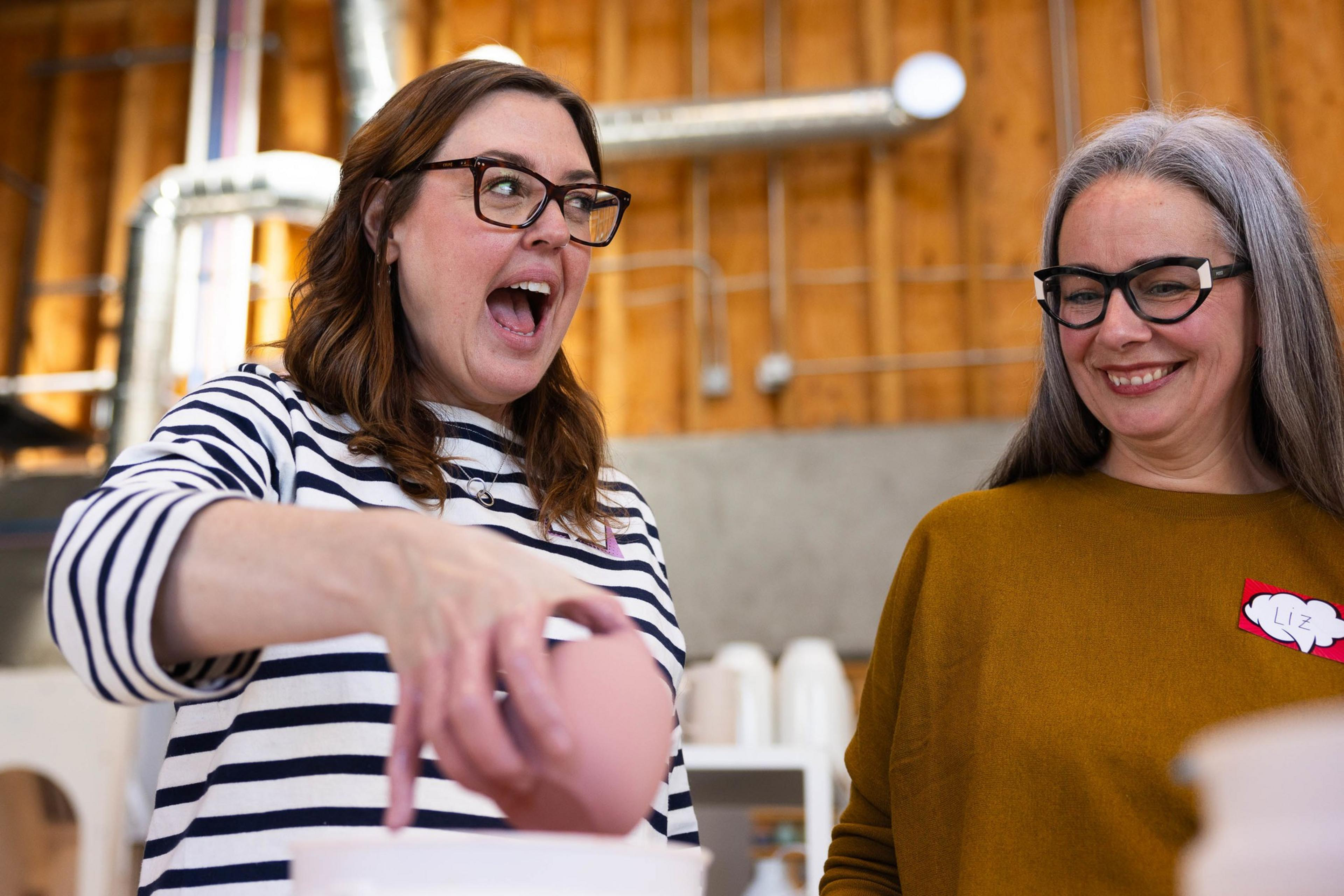 Two women smiling, one holding a bud vase, inside a room with exposed wooden beams.