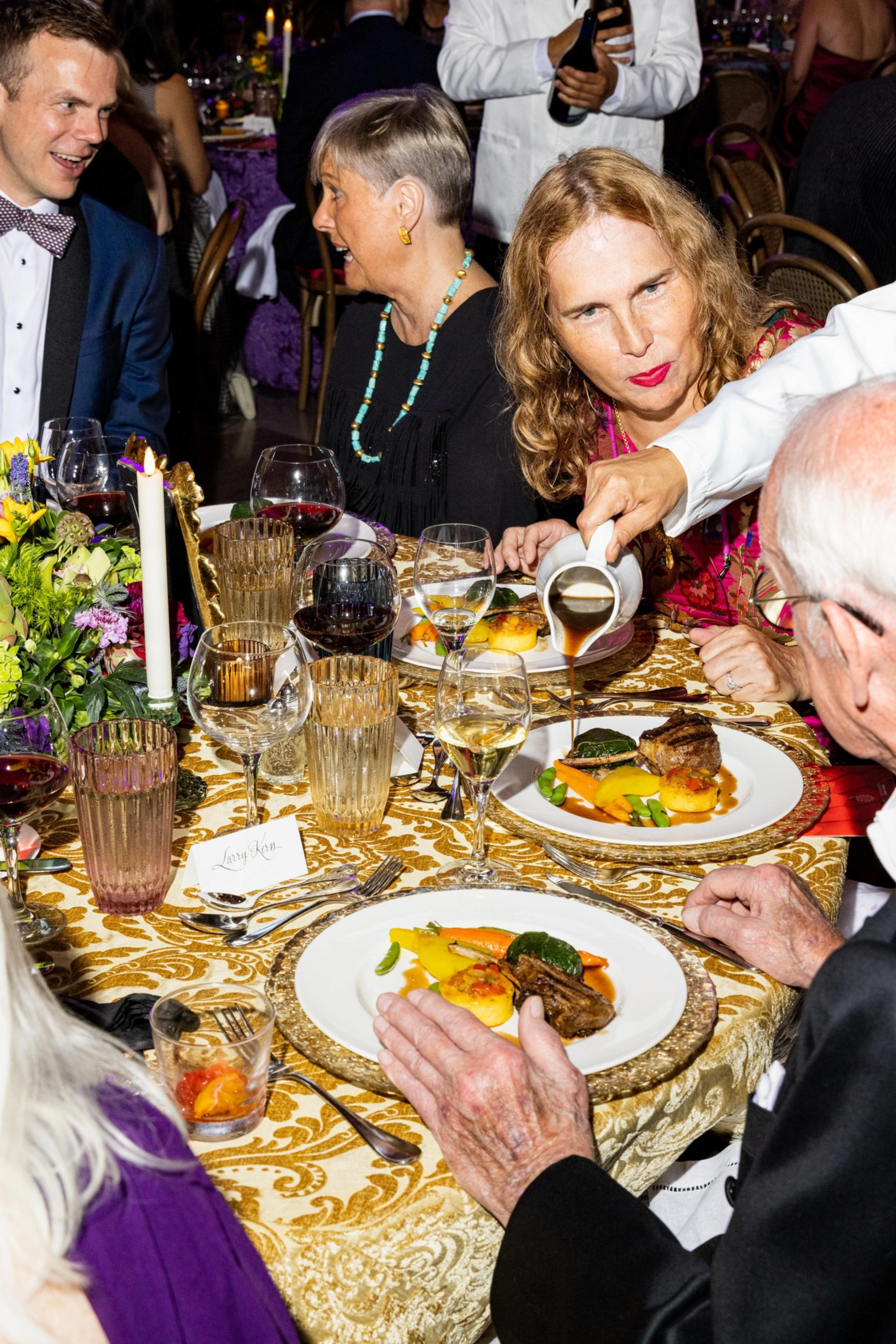 Four people sit around a table with ornate gold tablecloth, enjoying plated meals as a server pours sauce on one dish. Wine glasses and floral centerpiece are visible.