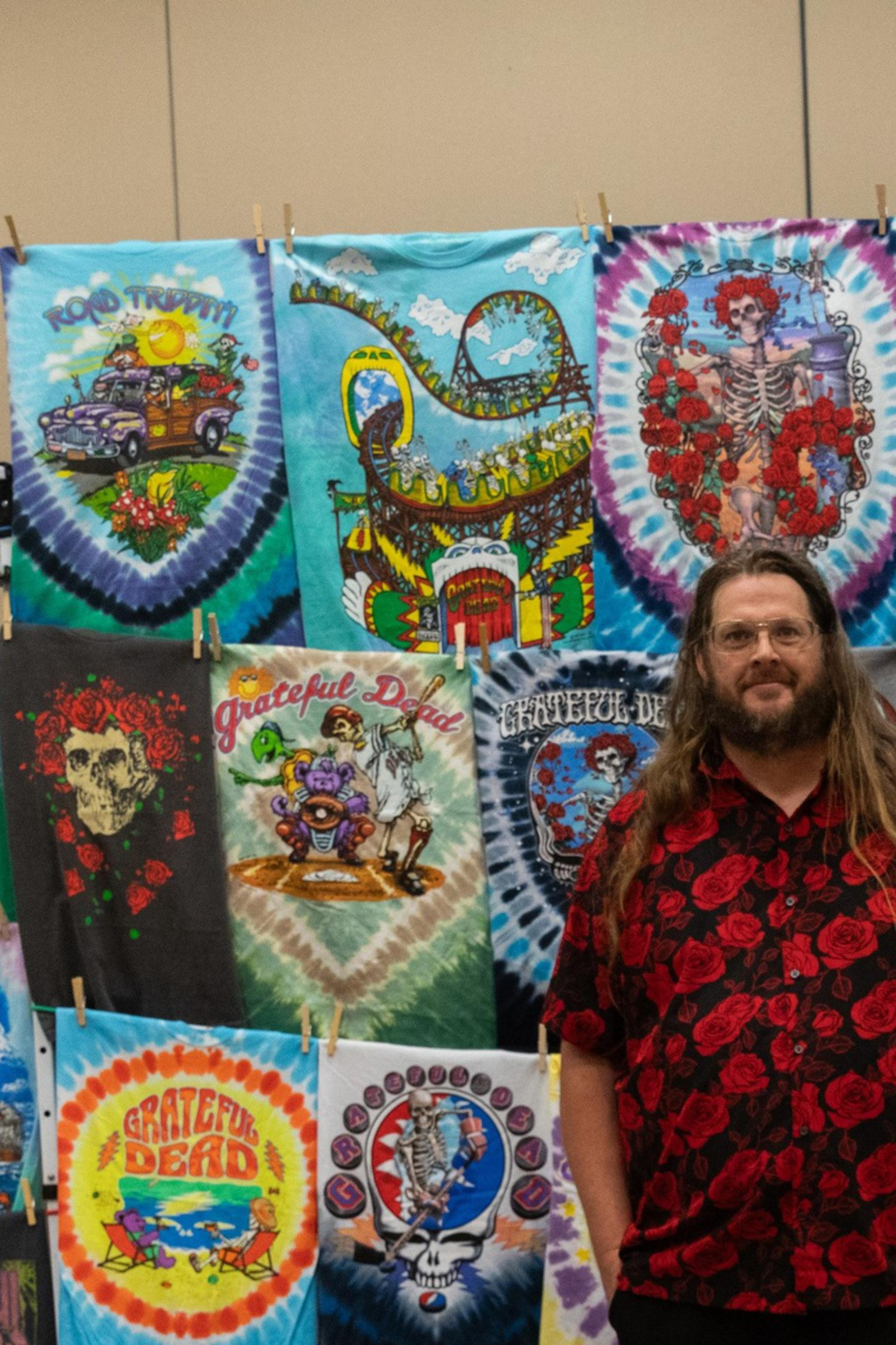A person stands in front of a colorful display of tie-dye and graphic T-shirts, with more shirts piled on the floor. They wear a red rose-patterned shirt and black shorts.