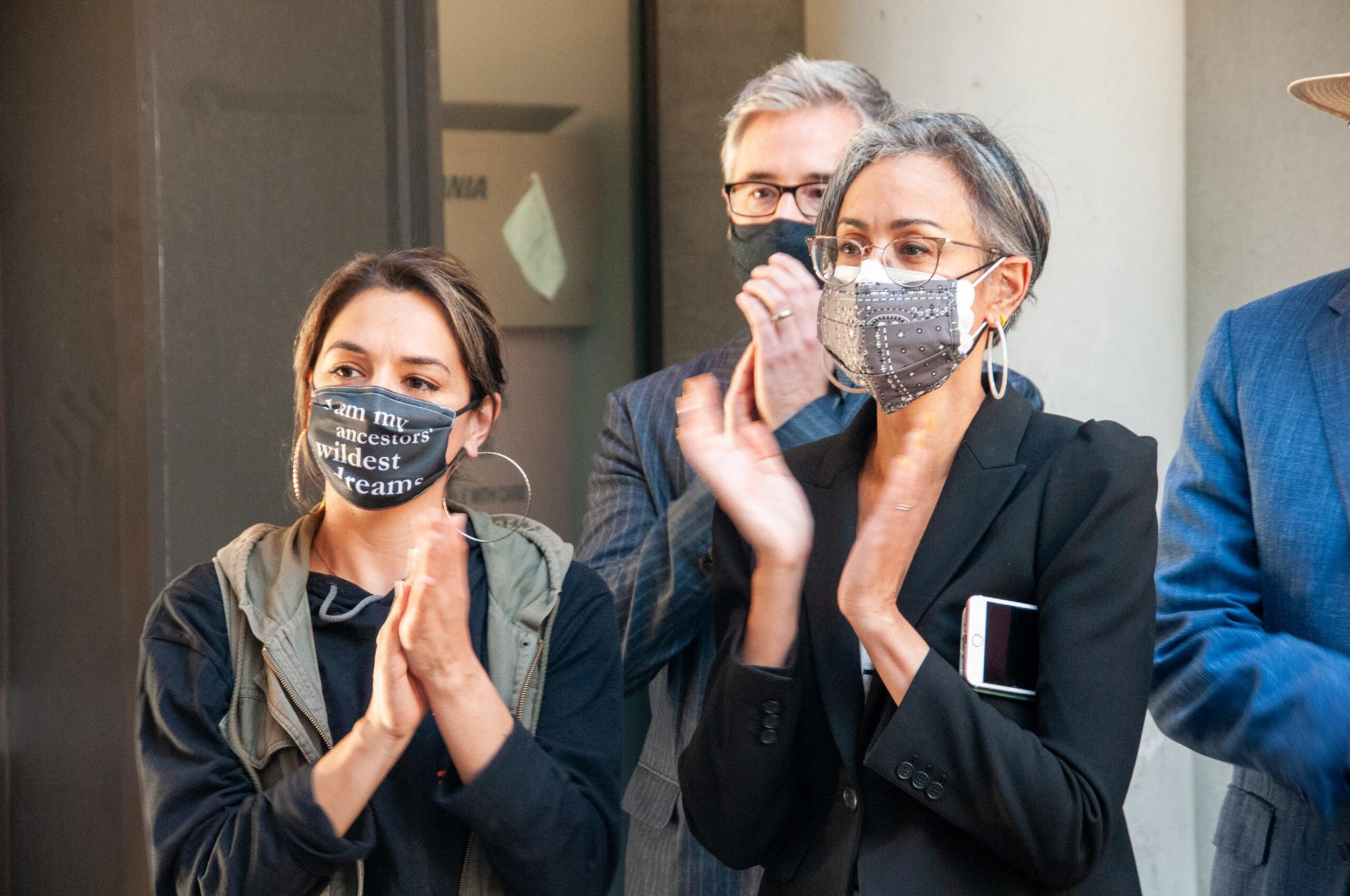Three people wearing face masks are clapping, two women in front and a man behind them, standing in a formal or public setting.
