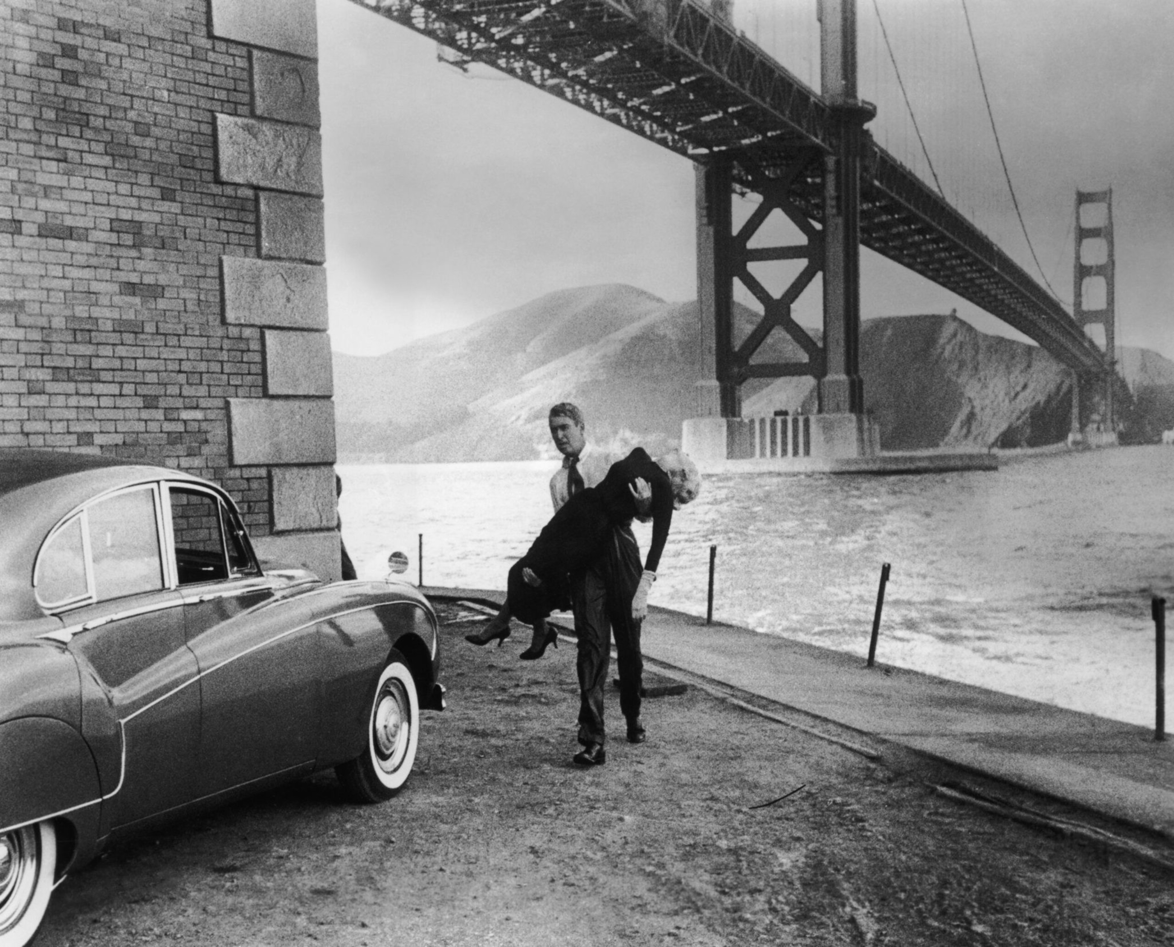 A man carries an unconscious woman near a classic car by the water, with the Golden Gate Bridge and hills in the background.