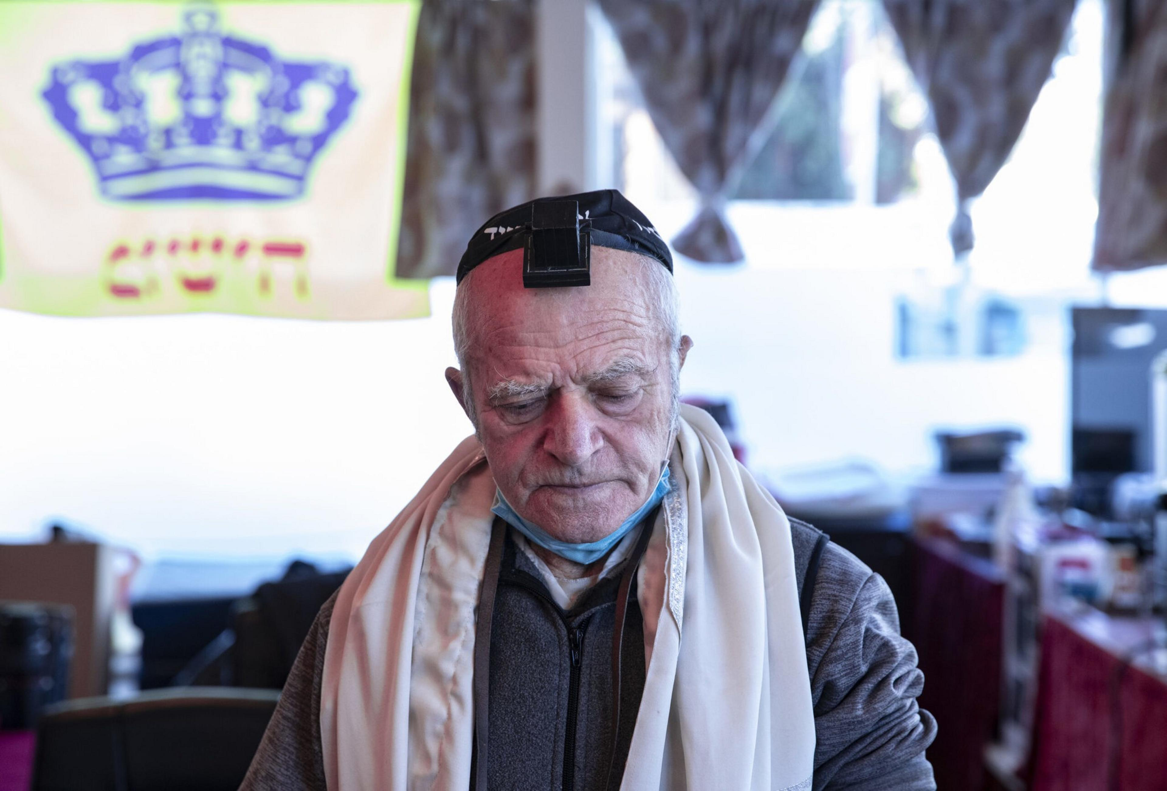 An elderly man wearing Jewish prayer garments, including tefillin on his forehead and a tallit, sits with eyes closed in a softly lit room.