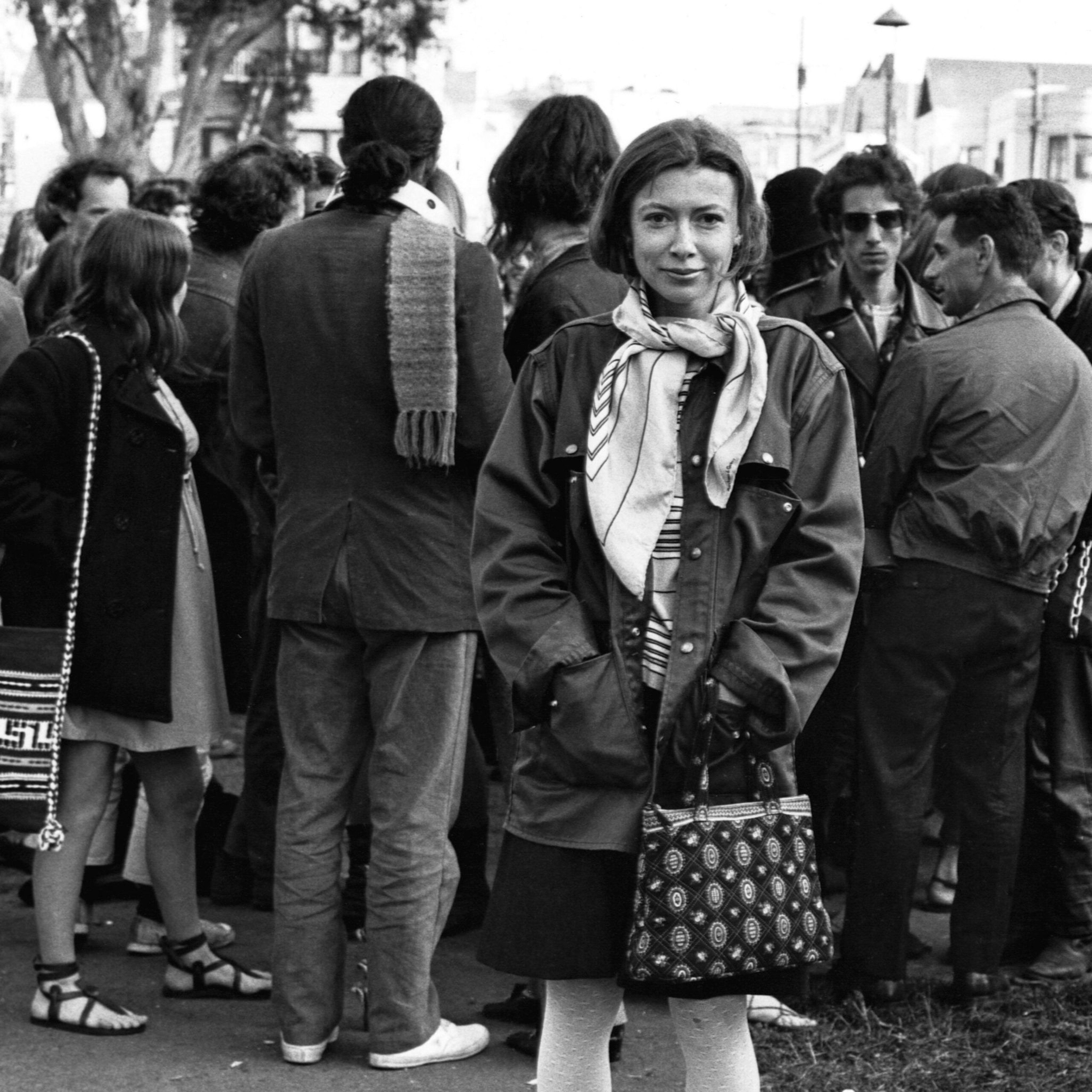 A woman with a scarf and patterned bag stands smiling in front of a crowd of casually dressed people, some facing away, in an outdoor setting.