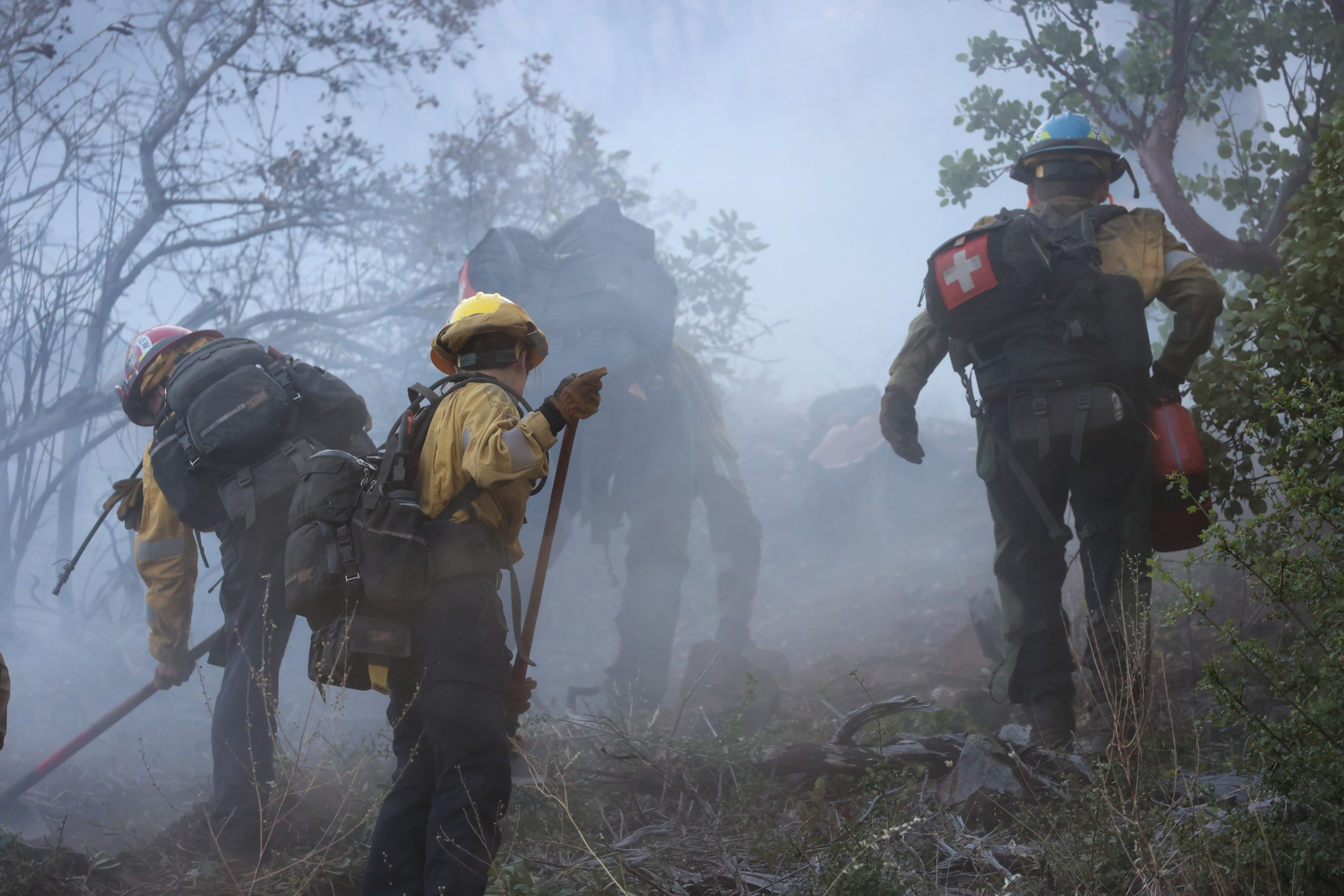 Firefighters in helmets and gear, carrying tools and backpacks, walk through a smoky, wooded area battling a wildfire.