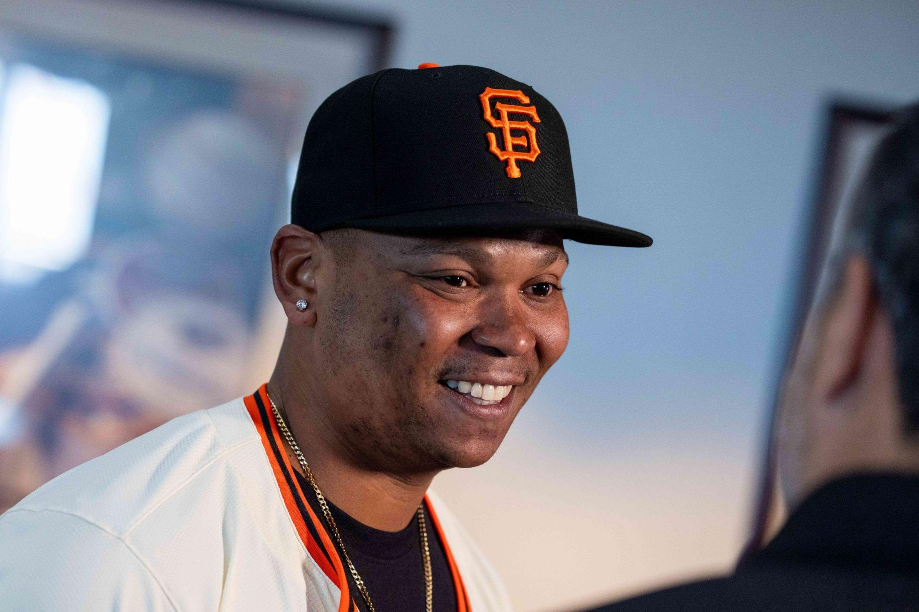 A person wearing a black cap with an orange logo smiles, facing another individual. They are dressed in a white jersey with black and orange trim.