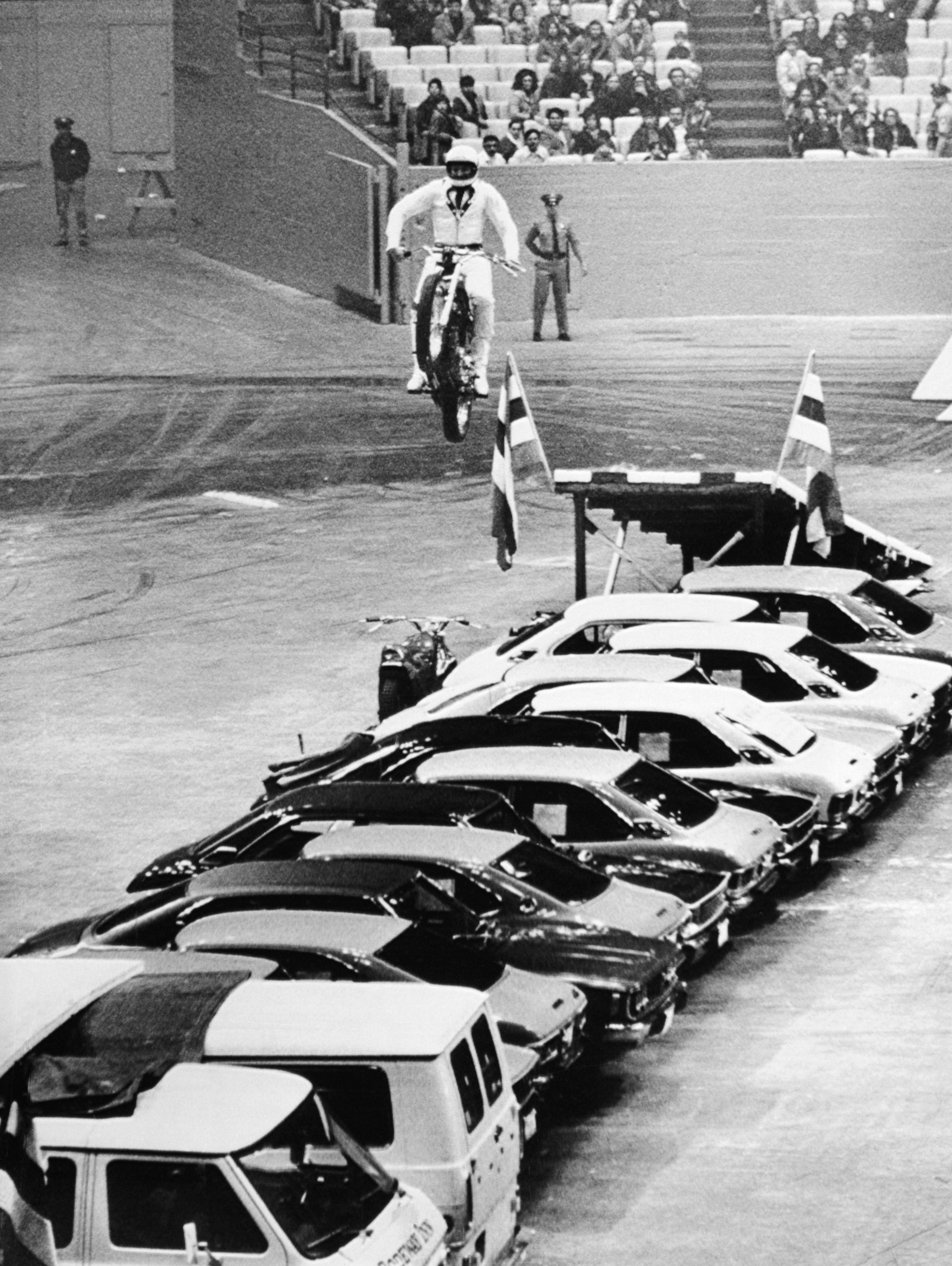 A motorcyclist in mid-air leaping over a long row of parked cars, in front of an audience.