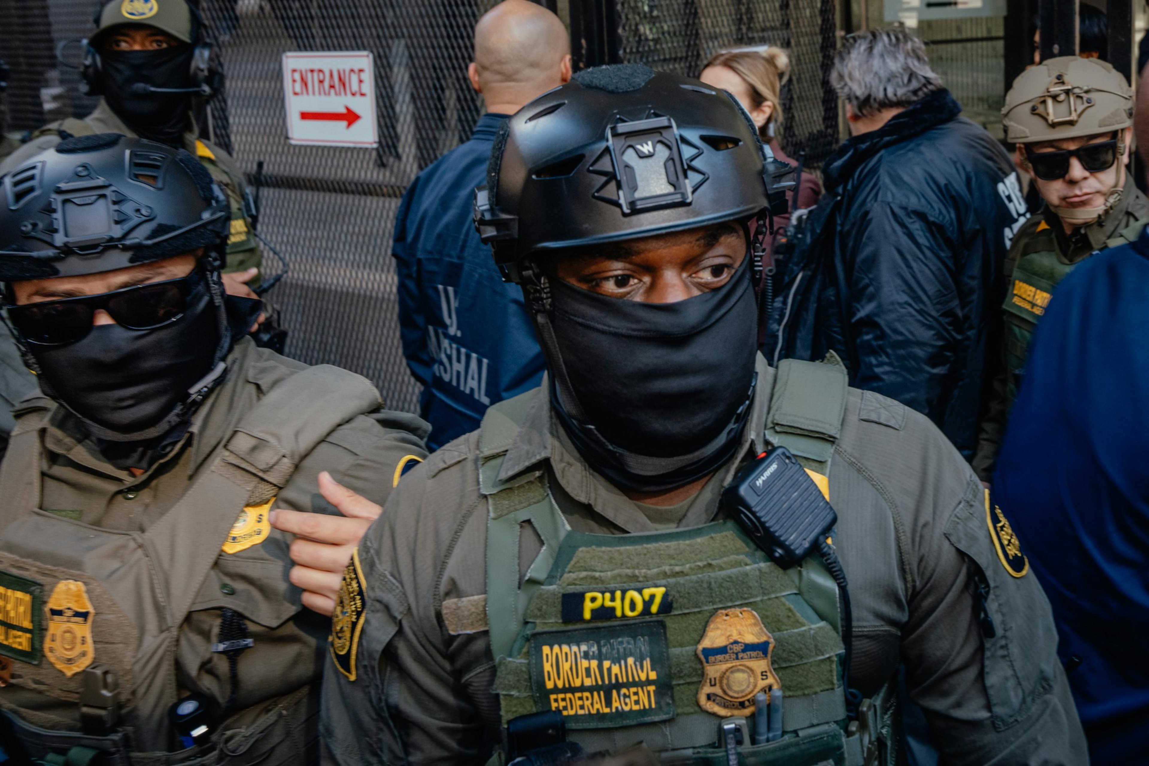 Several federal agents wearing tactical gear, helmets, and black face coverings stand together near a fence with an "ENTRANCE" sign.