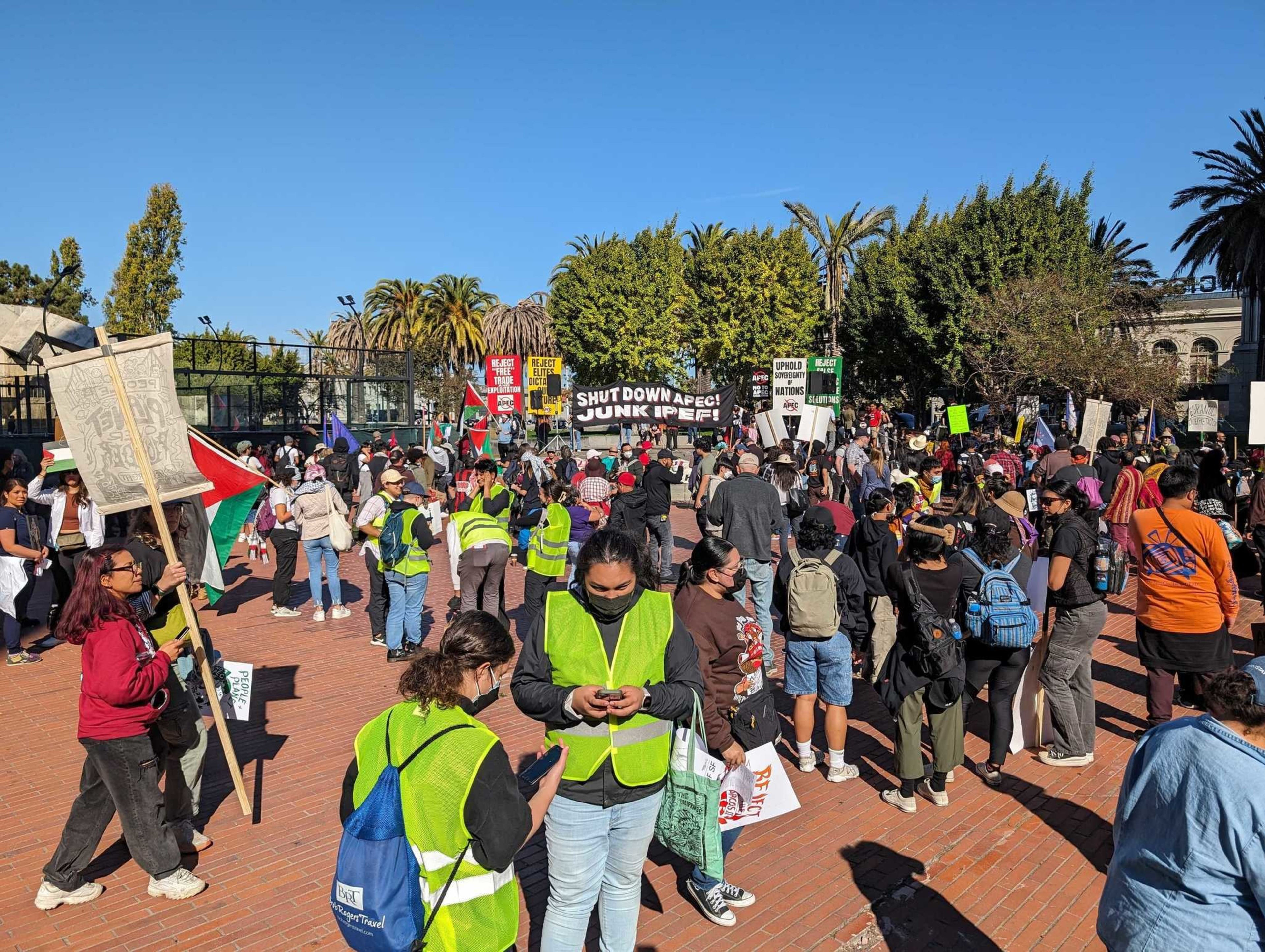 Hundreds of people in casual clothes, some holding signs, gather in a brick plaza on a sunny day.