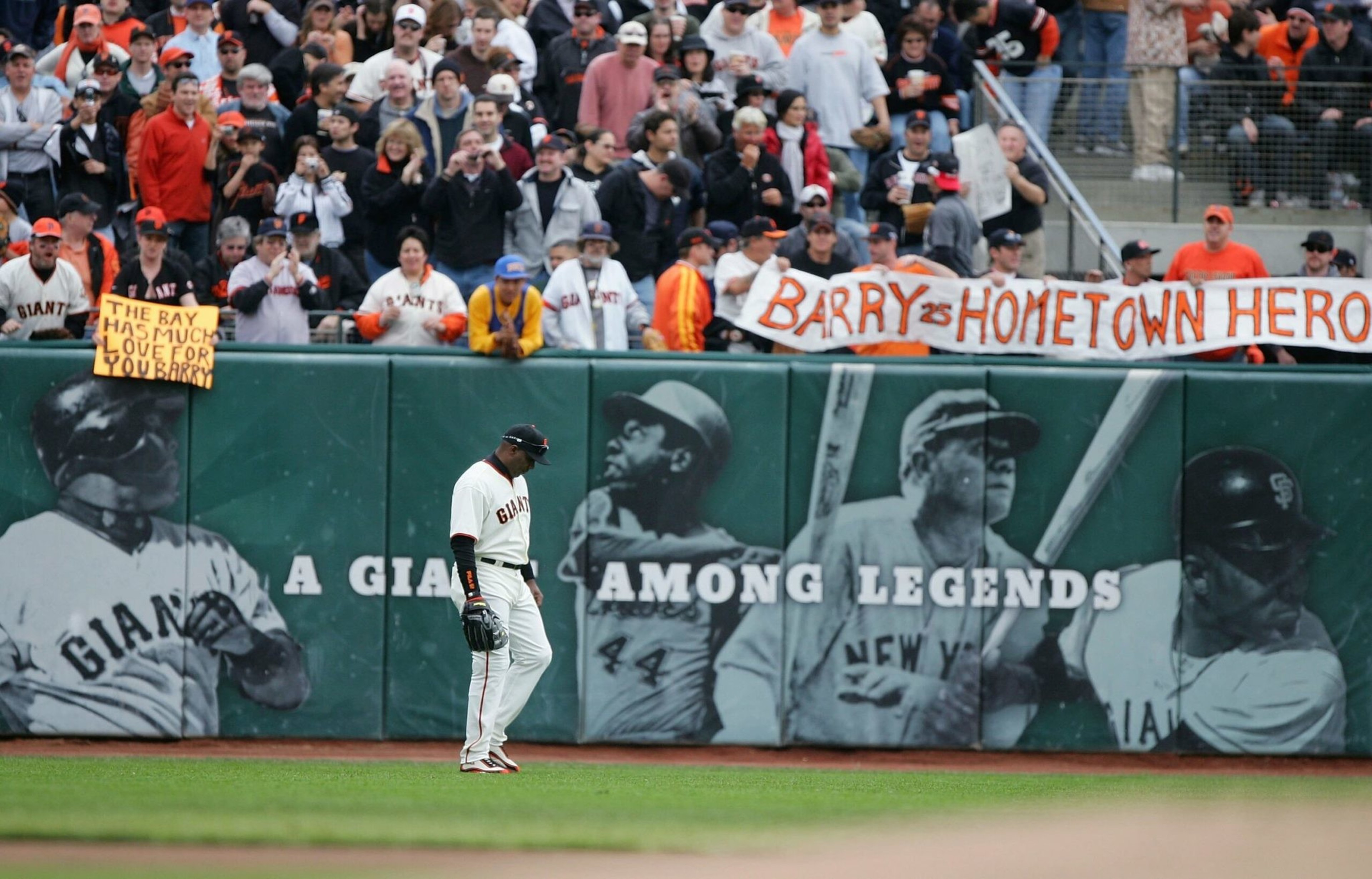 A Giants baseball player walks on the field with fans holding signs saying "The Bay has much love for you Barry" and "Barry is hometown hero" behind him.