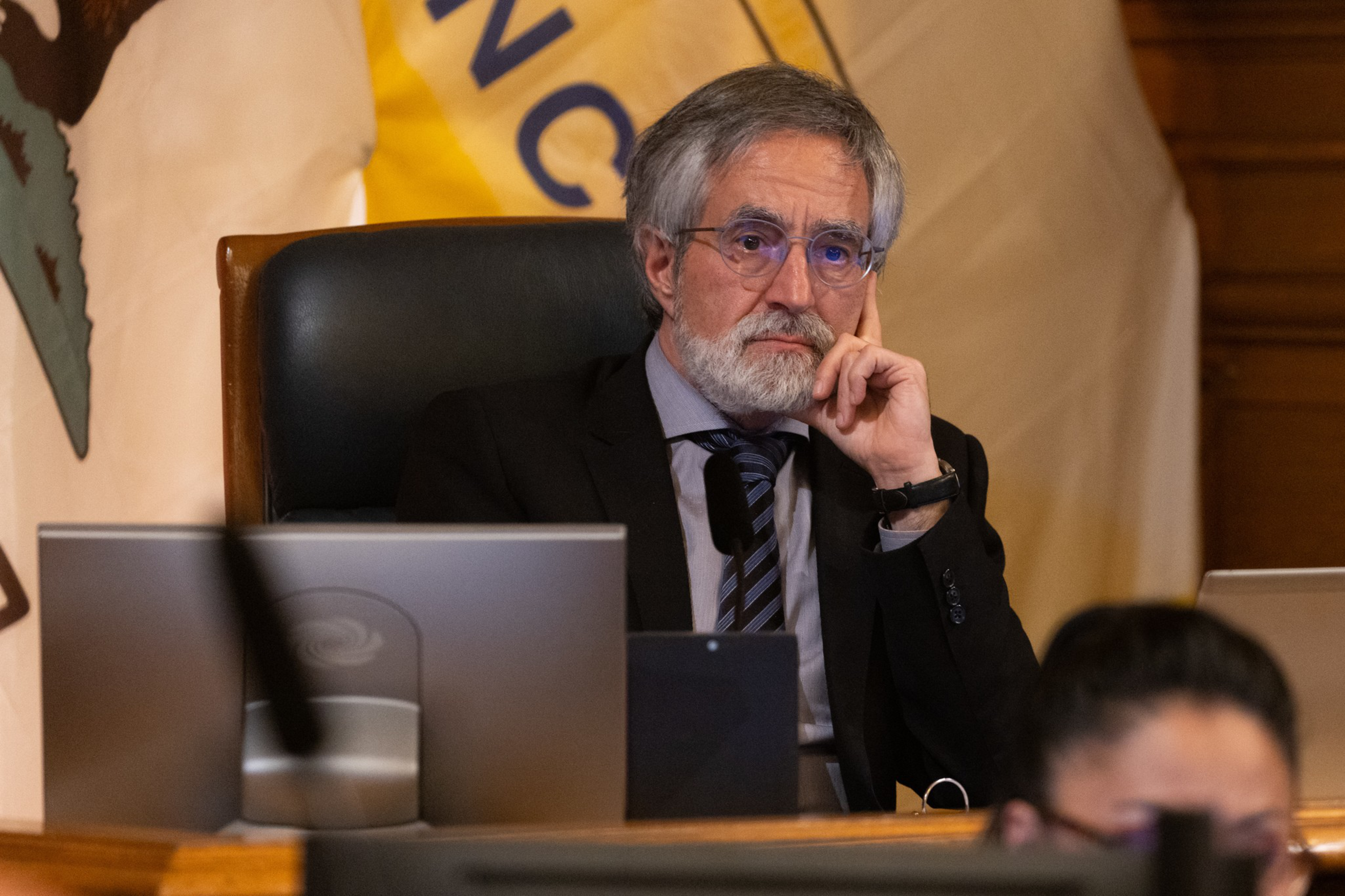 A pensive man with a beard and glasses, wearing a suit, rests his face on his hand, seated behind laptops.