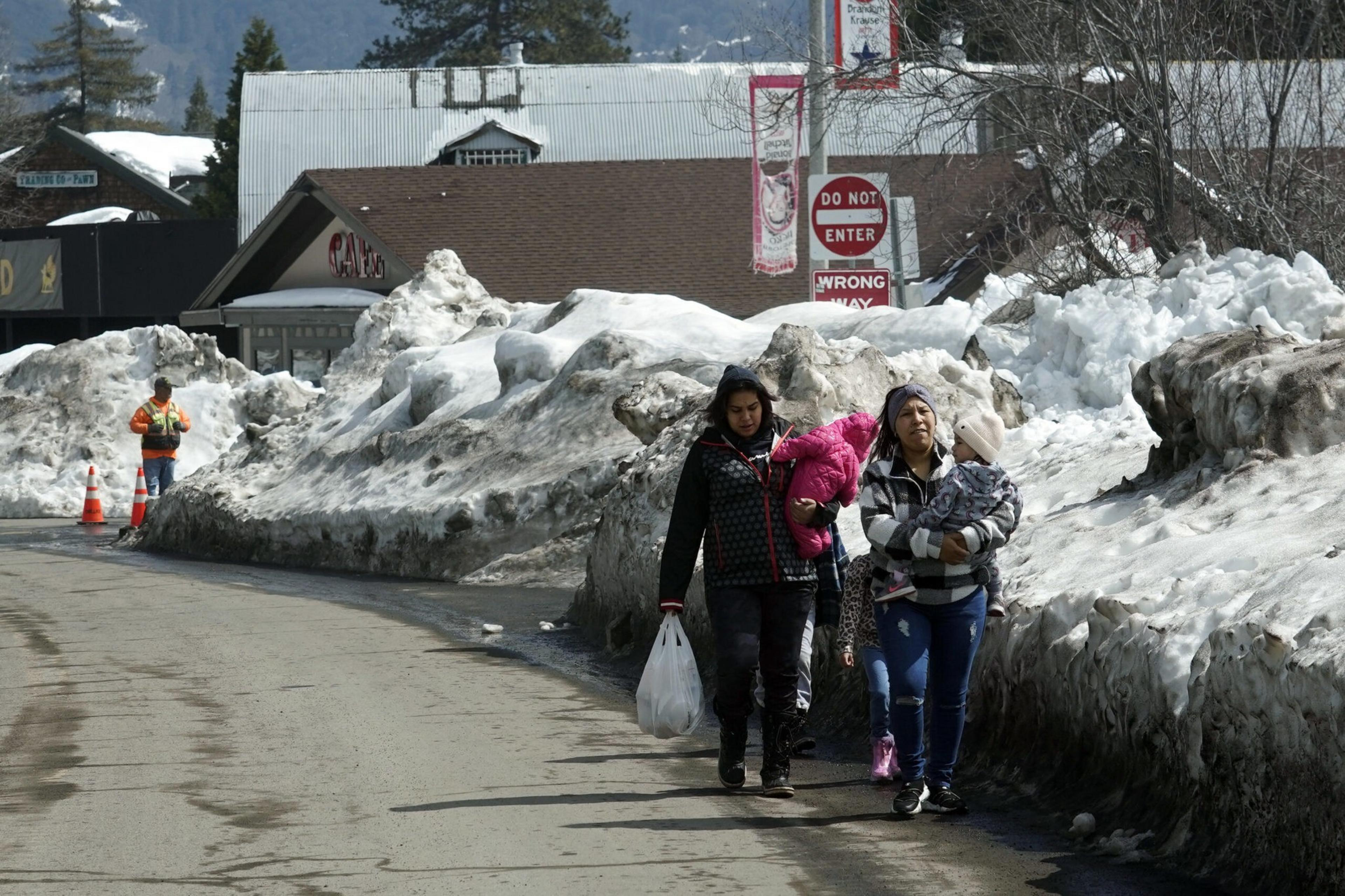 Two women walk along a snowy roadside, each carrying a child, with large dirty snow piles and a "Do Not Enter" sign in the background.