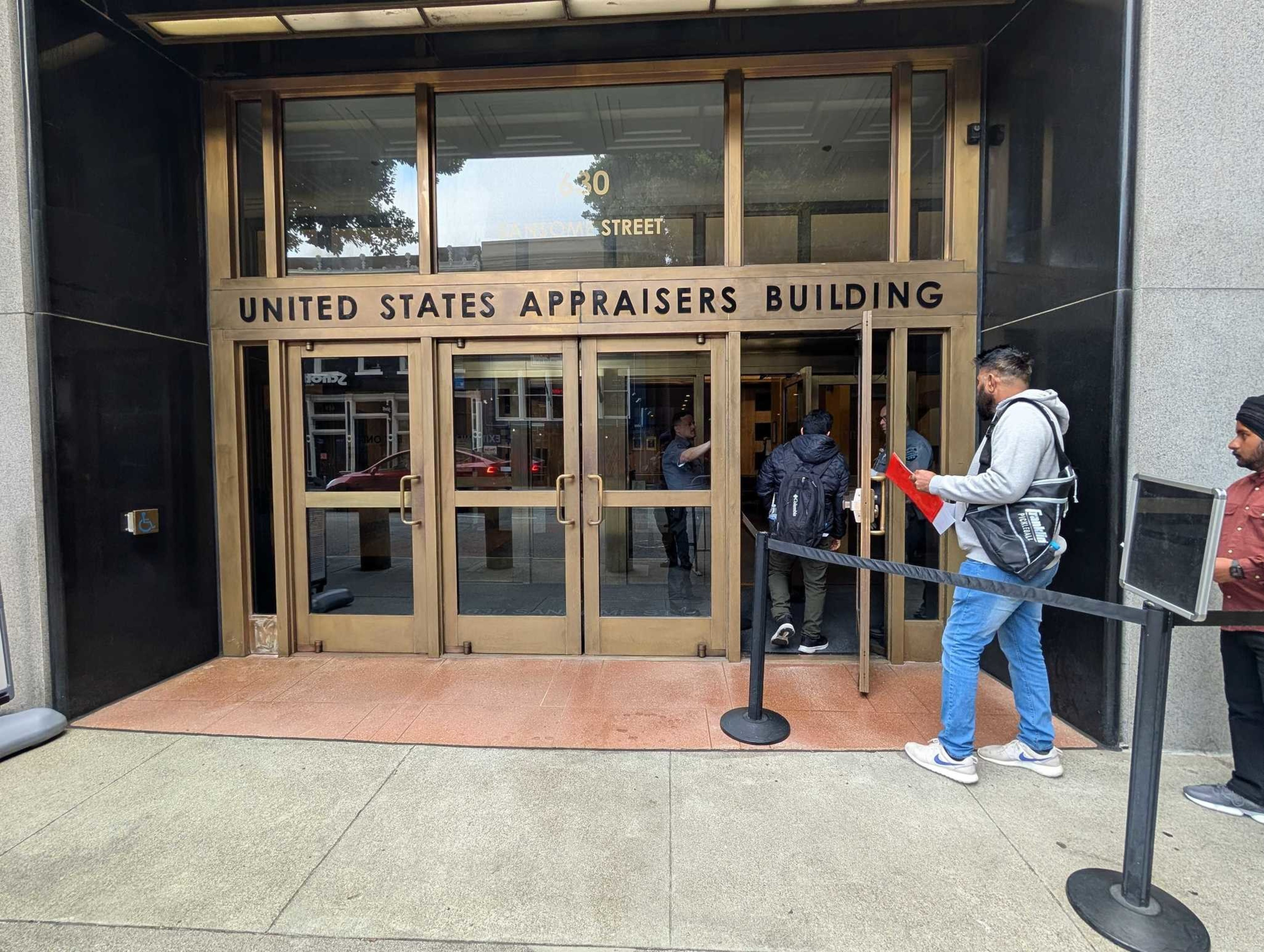 The image shows the entrance to the United States Appraisers Building with glass doors and people entering. A man with a backpack and red folder stands in line.