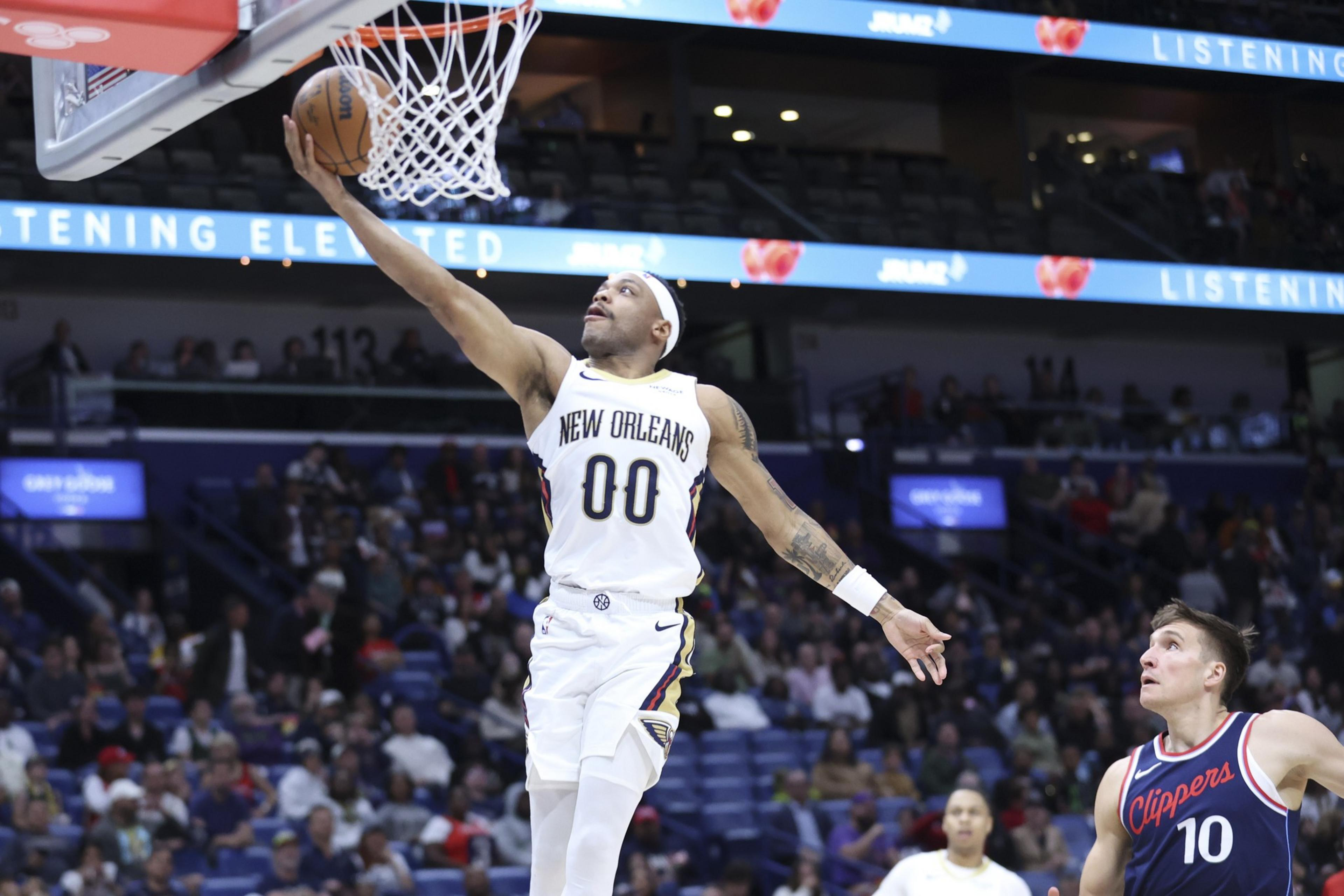 A basketball player from the New Orleans team is doing a layup while an opposing player from the Clippers watches. The crowd is visible in the background.