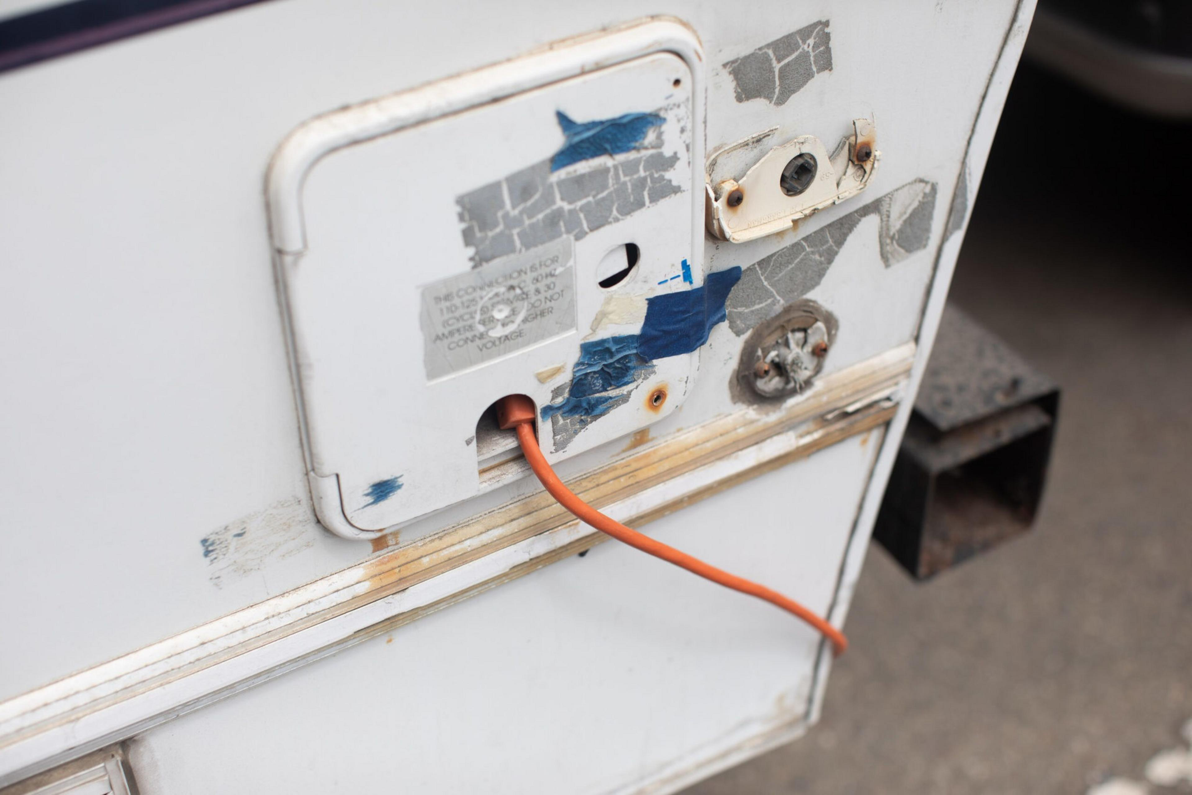A white metal panel with worn, peeling gray and blue tape has an orange cable plugged into a small rectangular opening at the bottom.