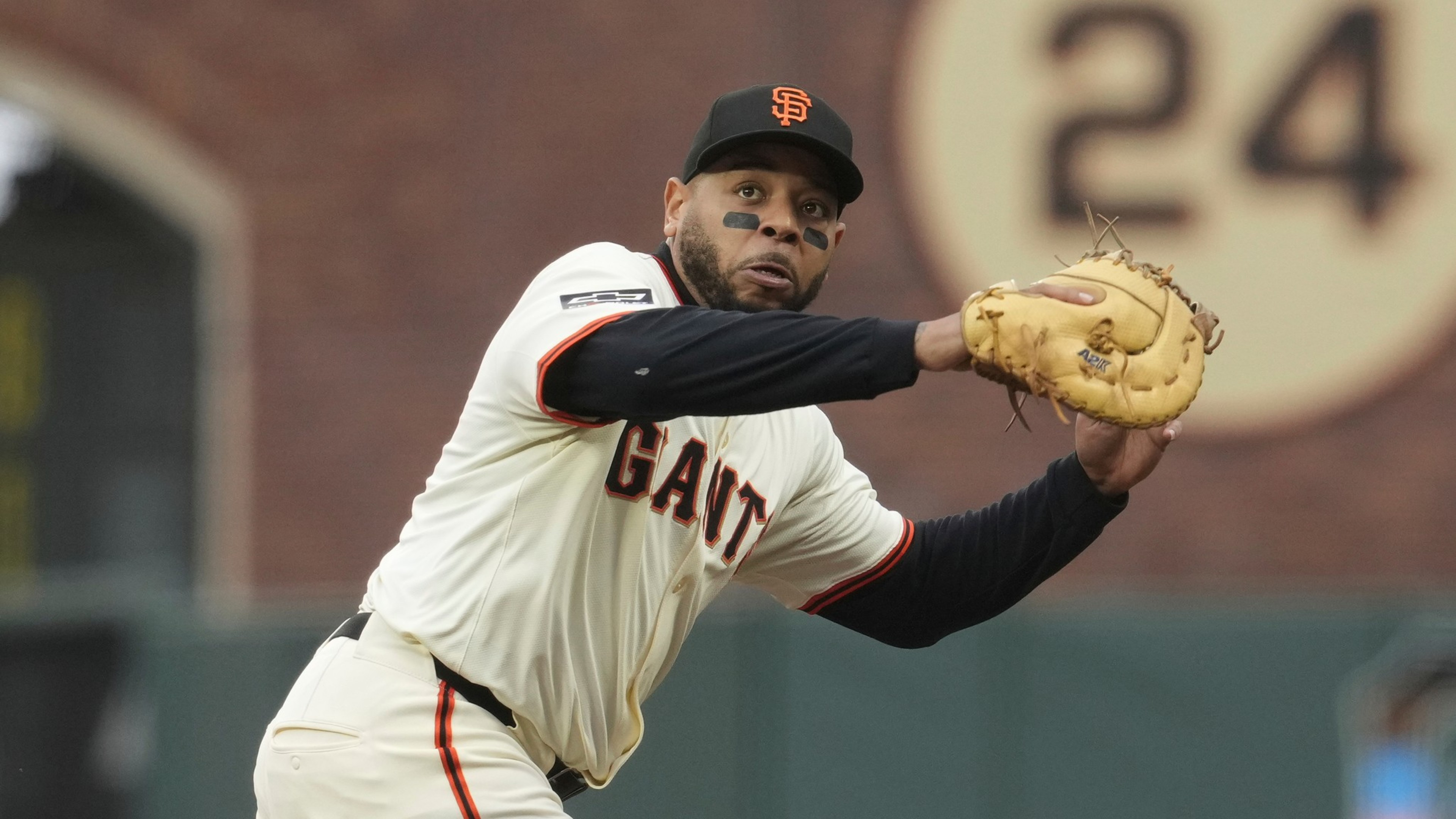 A baseball player wearing a Giants uniform is focused, mid-action, with a glove ready. Behind, a large number &quot;24&quot; is visible on the stadium wall.