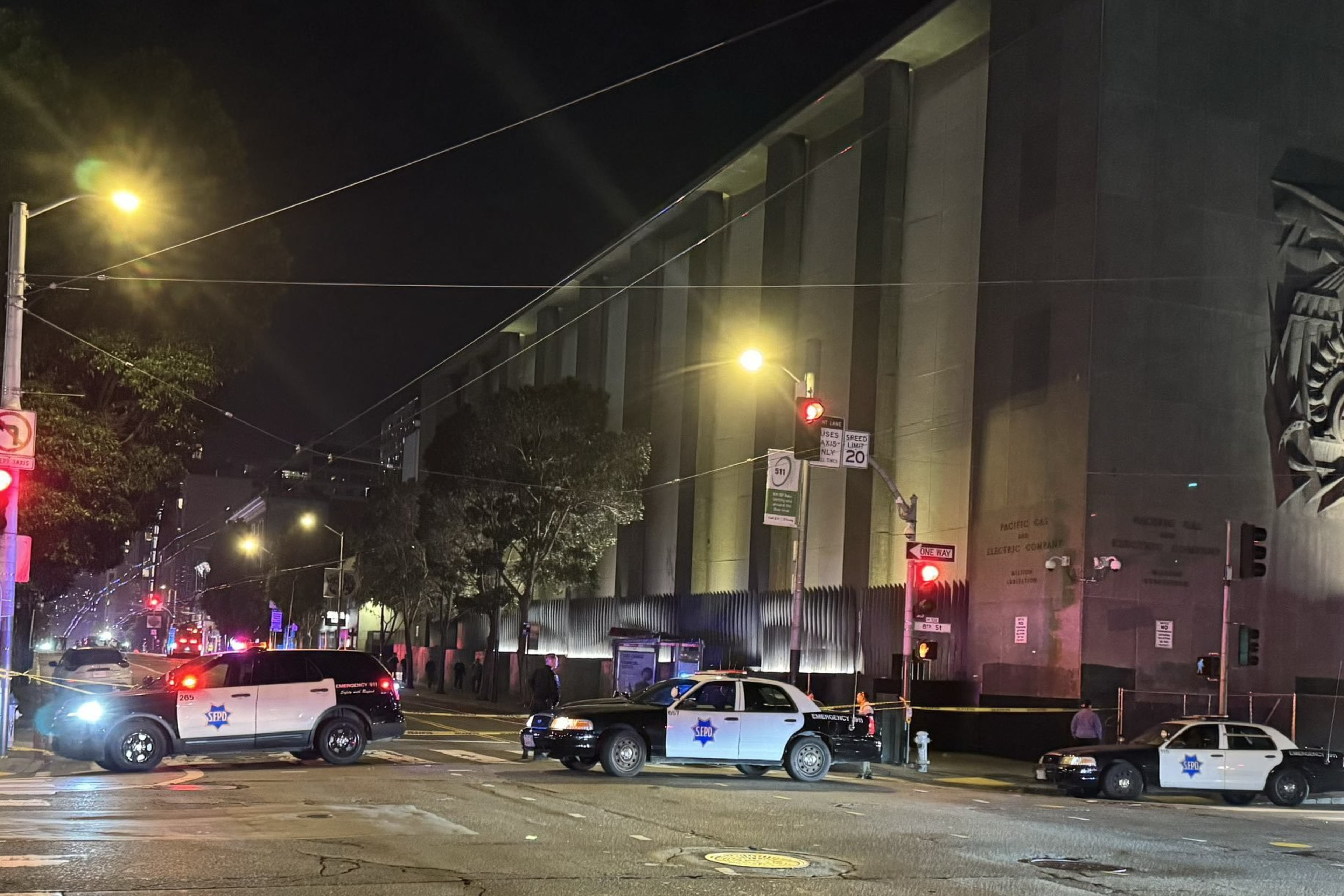 Three police cars block a street at night near a large building with sculpted art on the wall. Bright streetlights illuminate the scene.