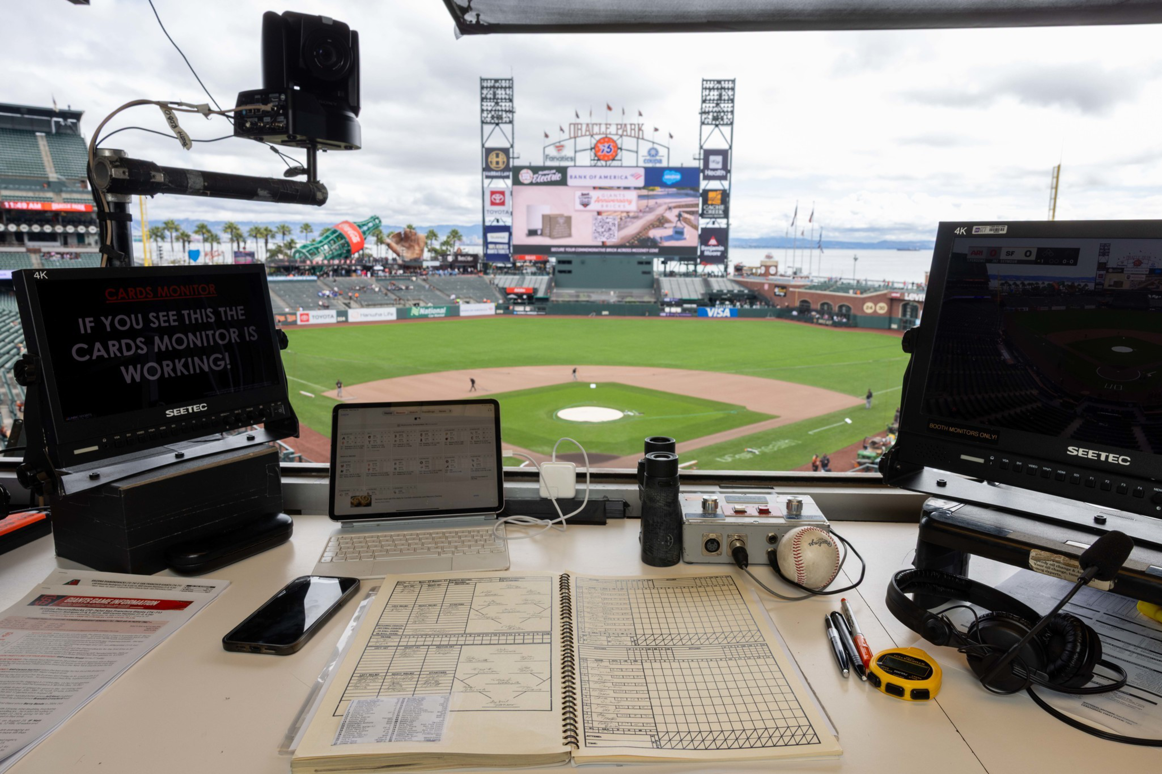A workspace with multiple monitors, a camera, and audio equipment overlooks a baseball field through a large window.