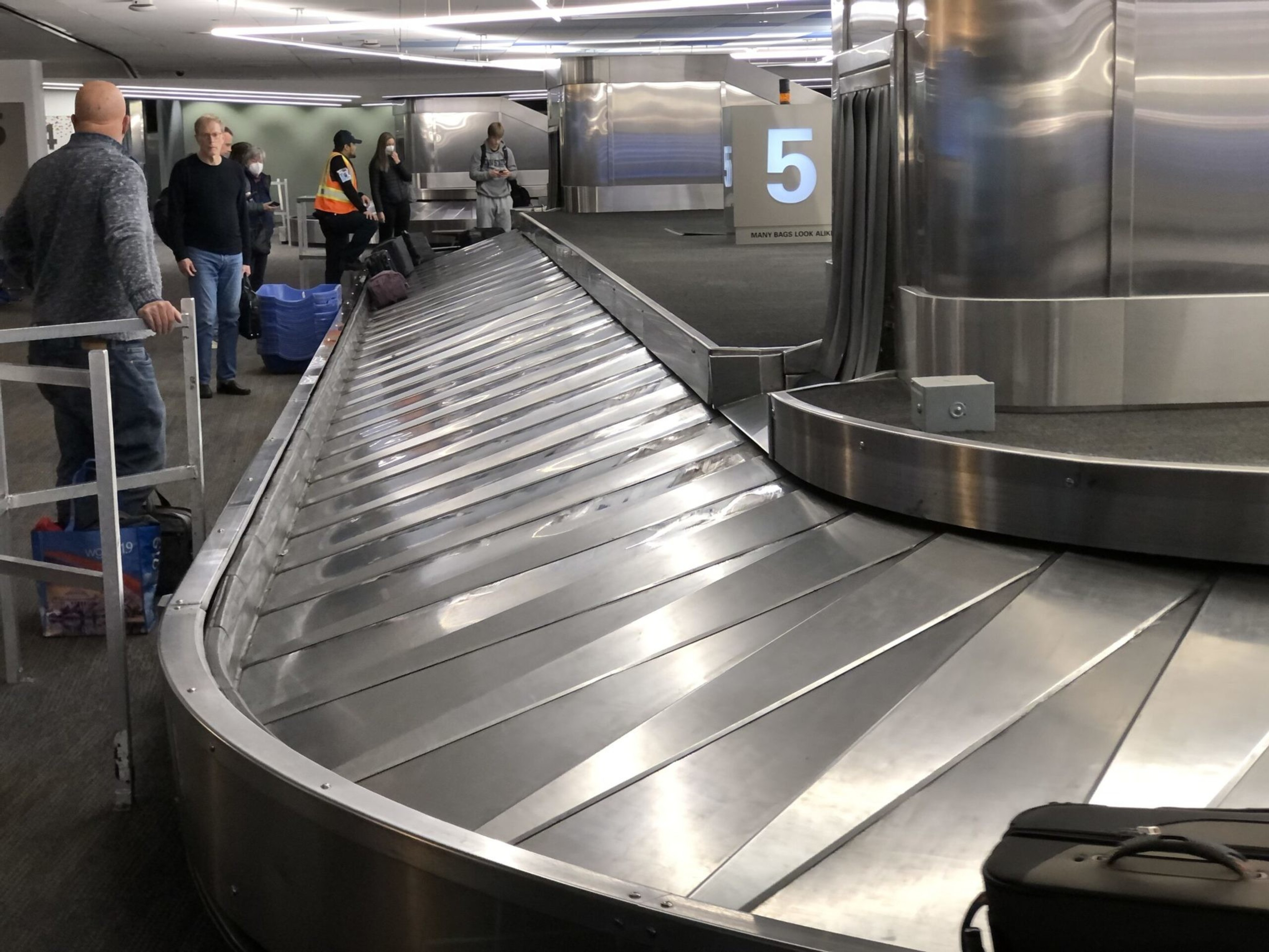 A mostly empty airport baggage claim carousel with a few people waiting and one suitcase visible near the end.