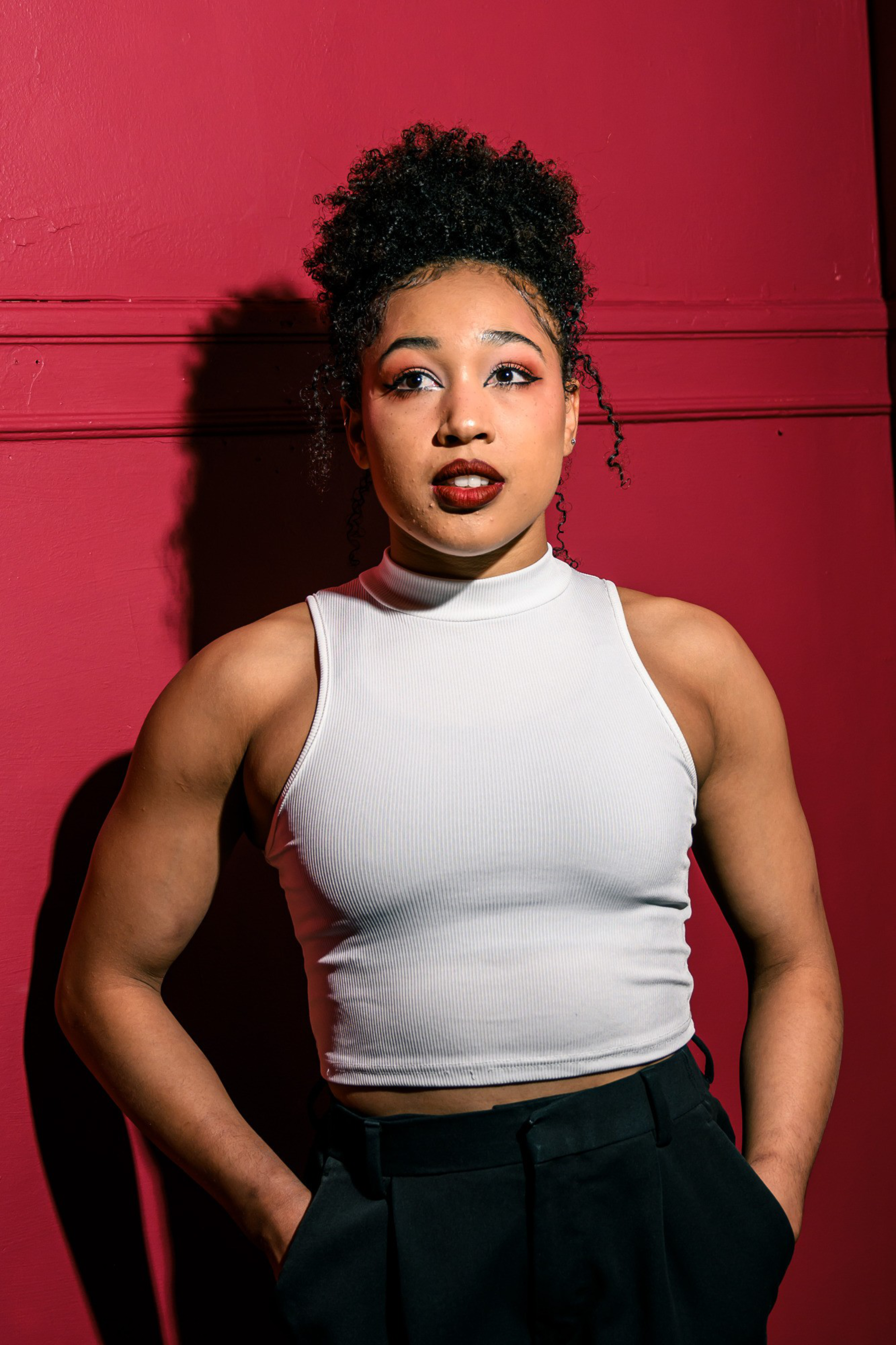 A person with curly hair stands against a red wall, wearing a white sleeveless top and black pants. Their expression is thoughtful, looking slightly upward.
