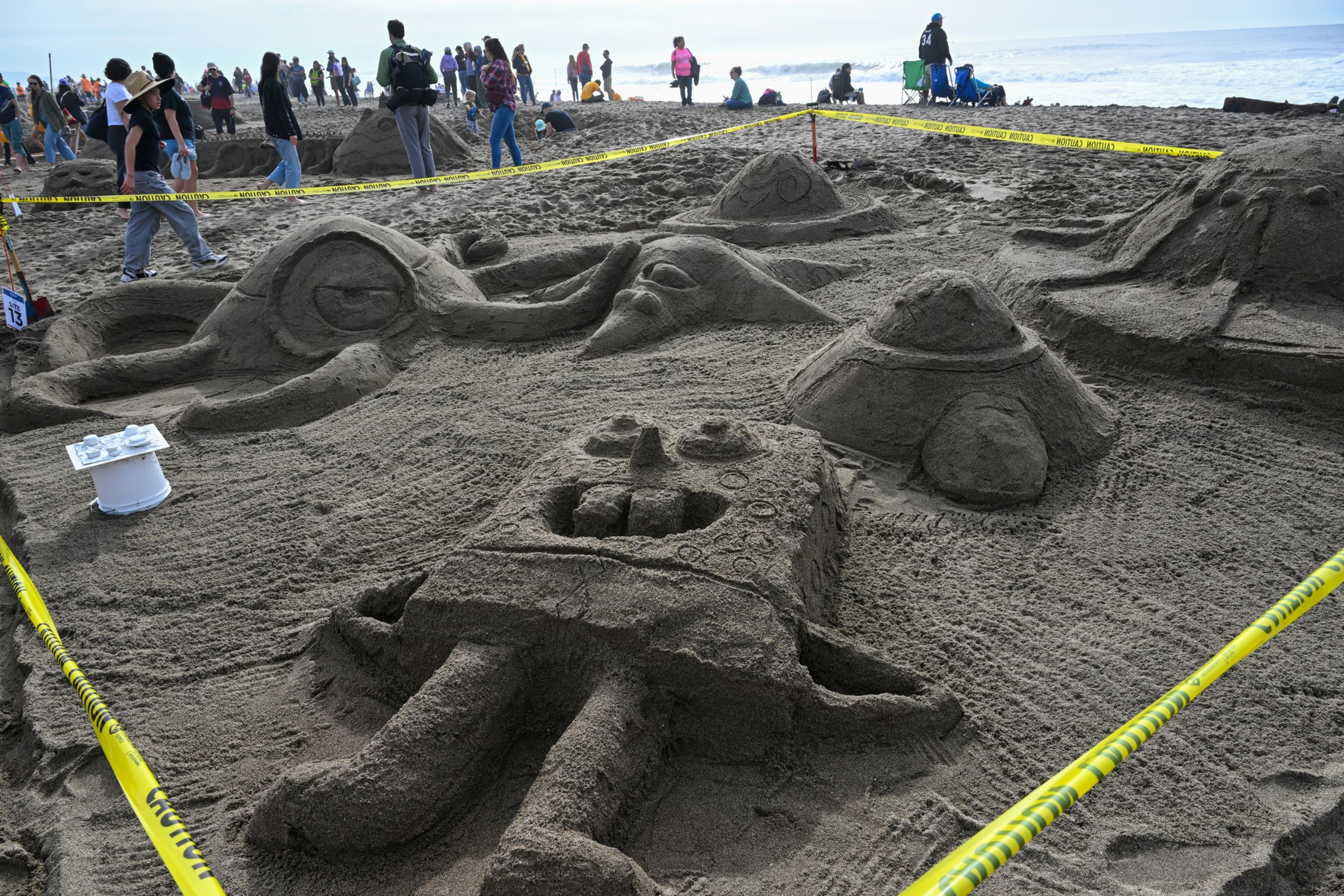 A detailed sand sculpture of SpongeBob SquarePants and other characters is roped off on a beach, with people walking and observing in the background.