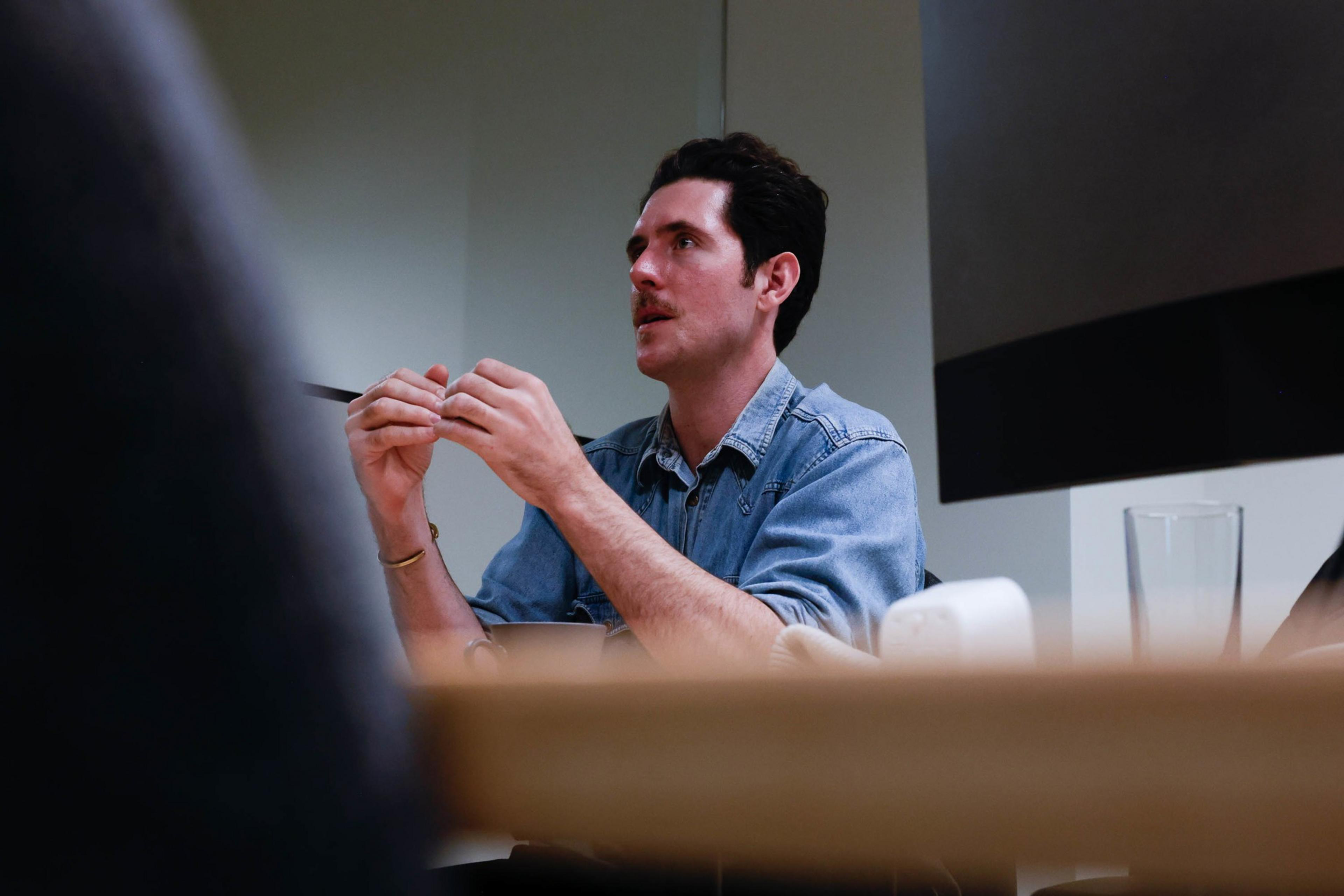 A man in a denim shirt is sitting at a table, looking up and holding a pen, with glasses and a water glass nearby.