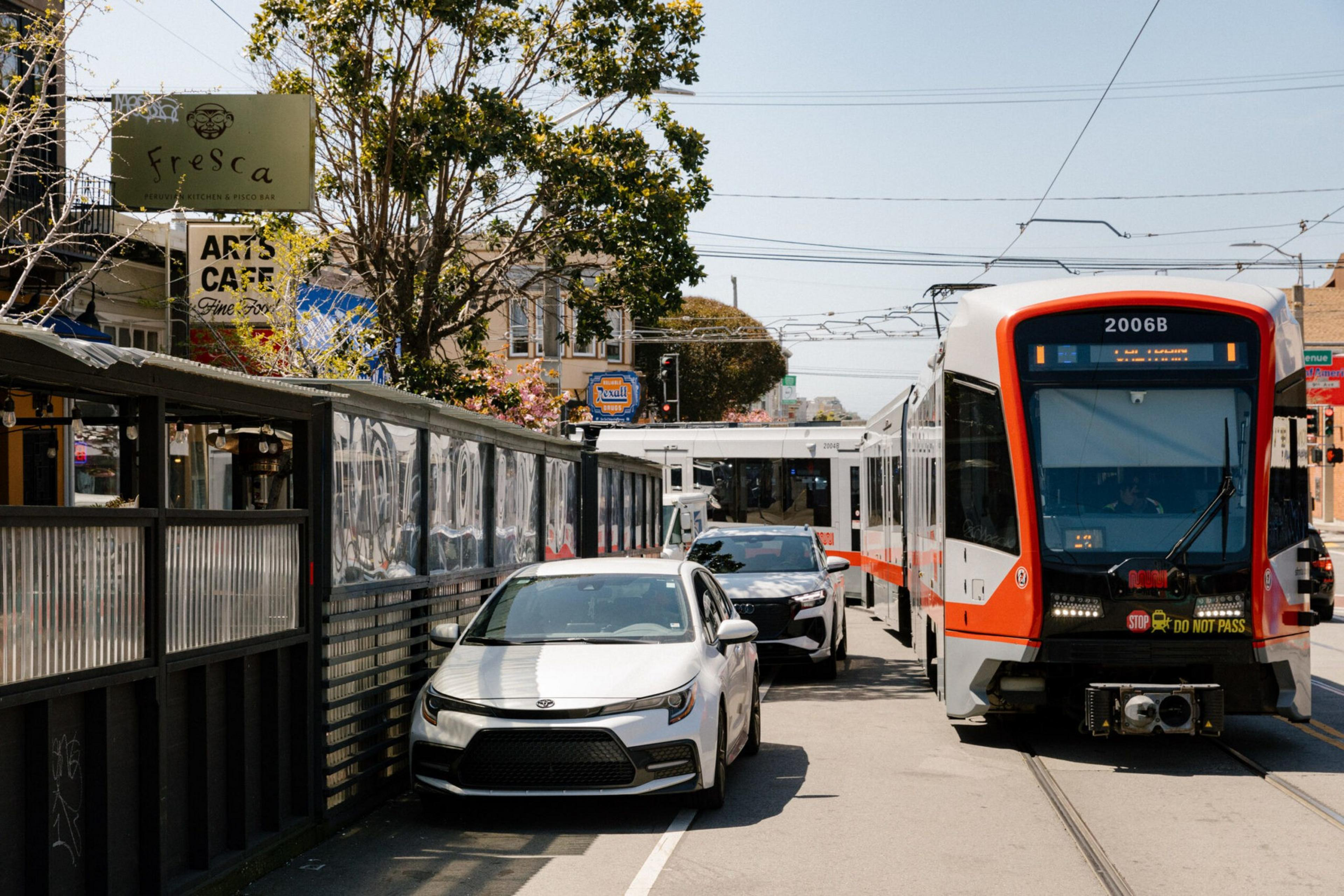 A white car and a black car are parked beside a streetcar with orange and white colors on city tracks, with outdoor cafe seating and trees nearby.
