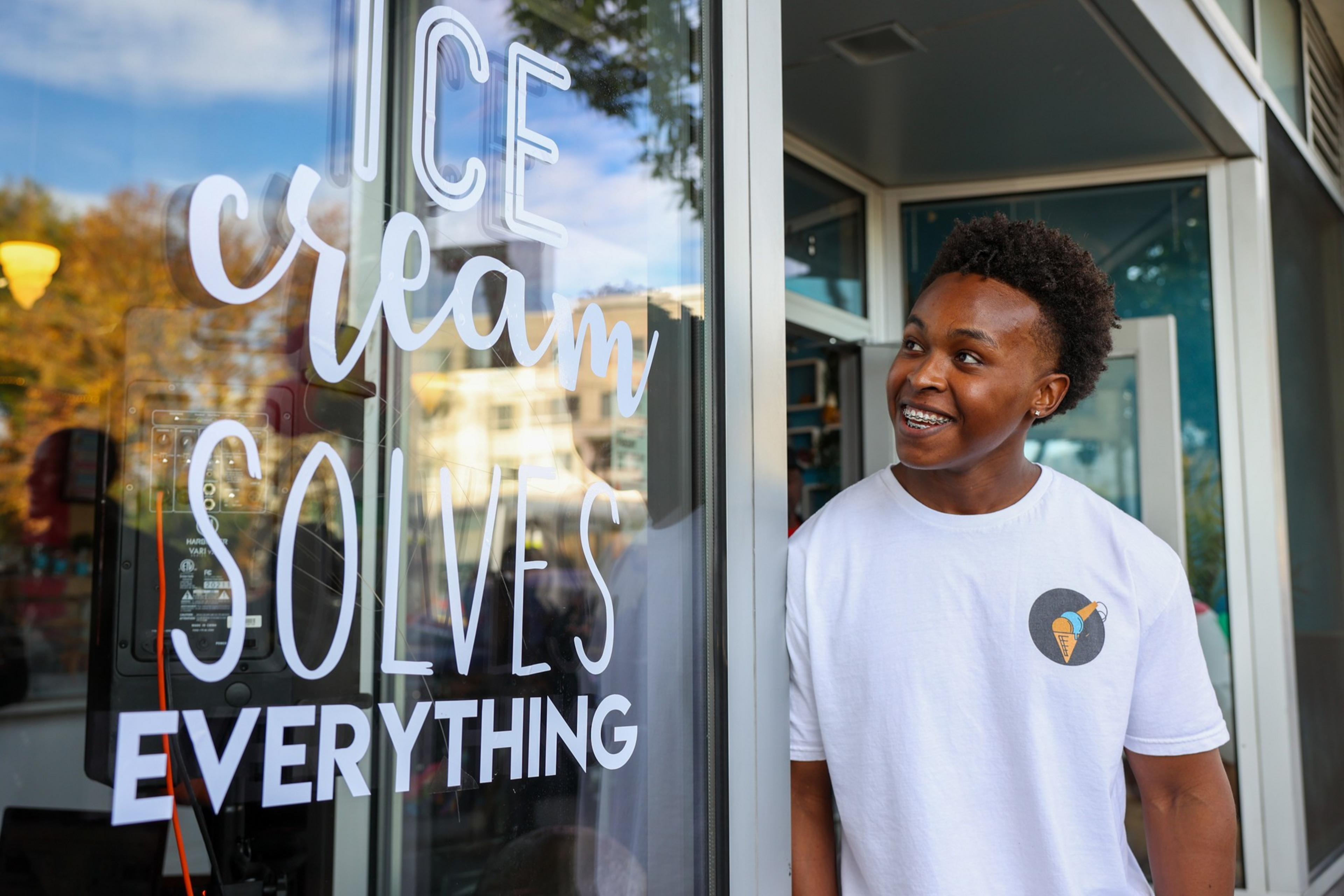 A smiling person with short curly hair and braces stands in a doorway next to a glass door with white text that reads, "Ice cream solves everything."