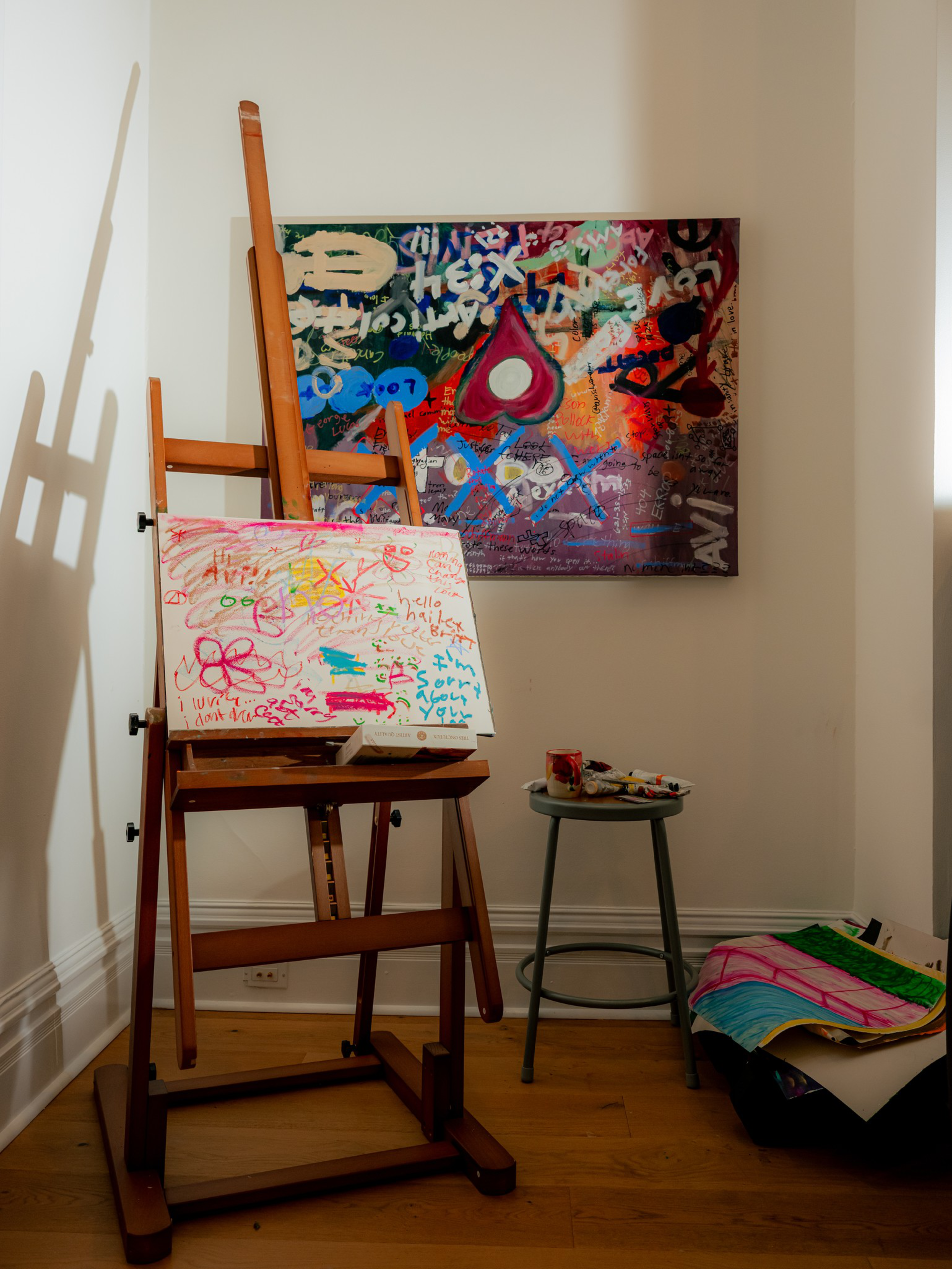 An artist’s wooden easel holds a colorful canvas with scribbles and drawings, next to a small stool with paint supplies and a vibrant abstract painting on the wall.