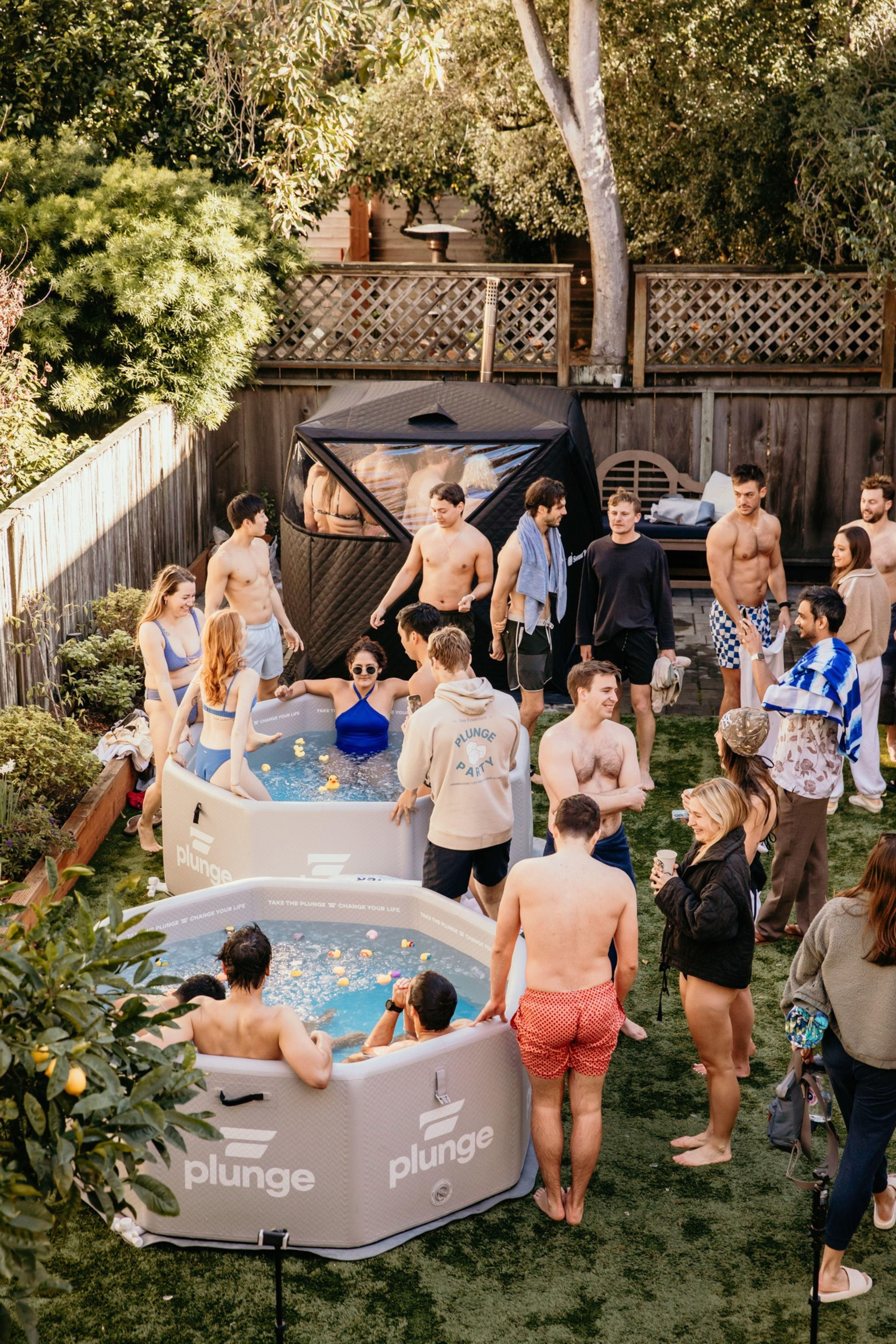 The image shows a lively backyard scene with people gathered around inflatable plunge pools, some dressed in swimsuits, socializing and enjoying a sunny day.