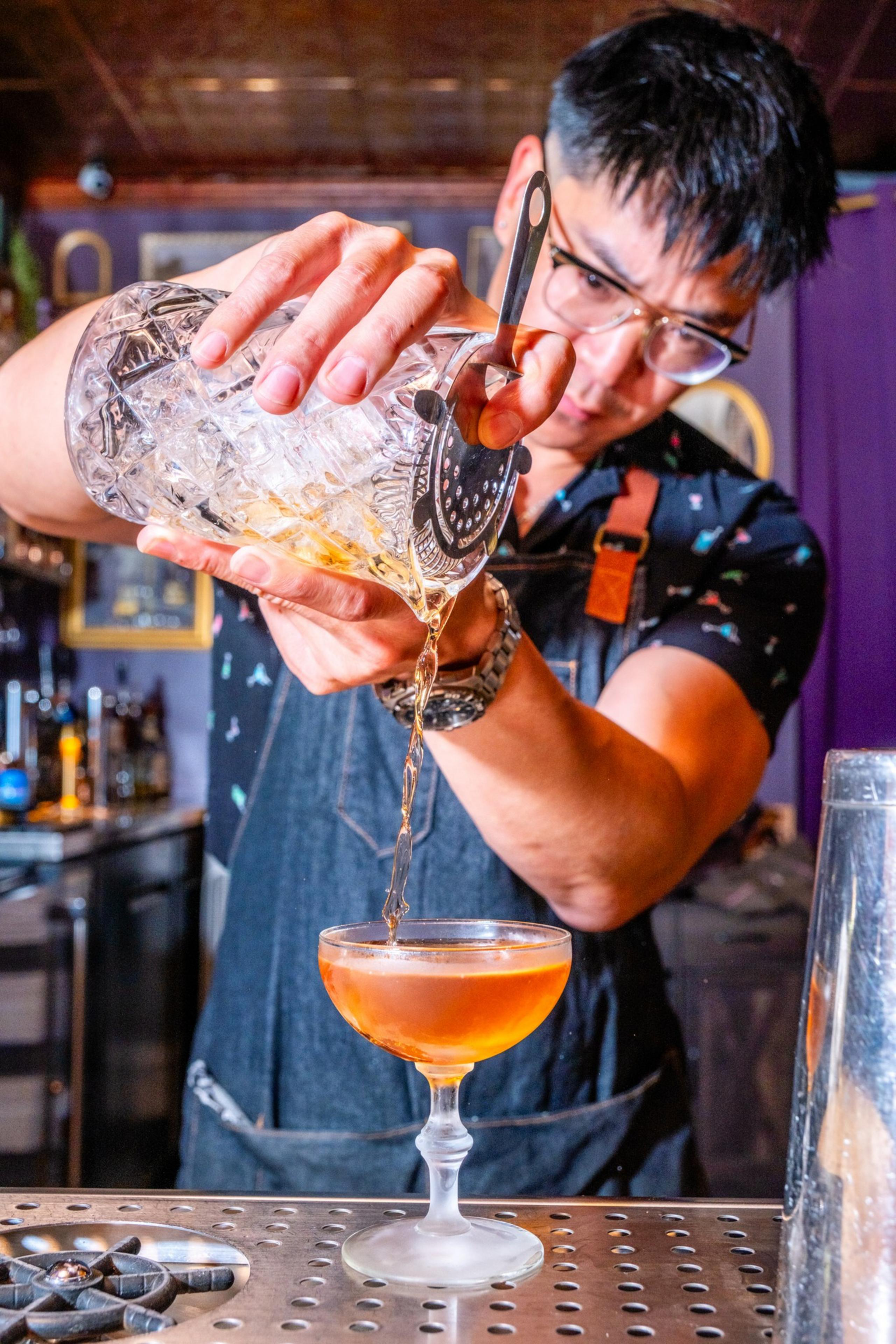 A bartender pours a golden cocktail into a glass, using a strainer over a mixing glass filled with ice. He wears a dark apron and shirt, focusing intently.