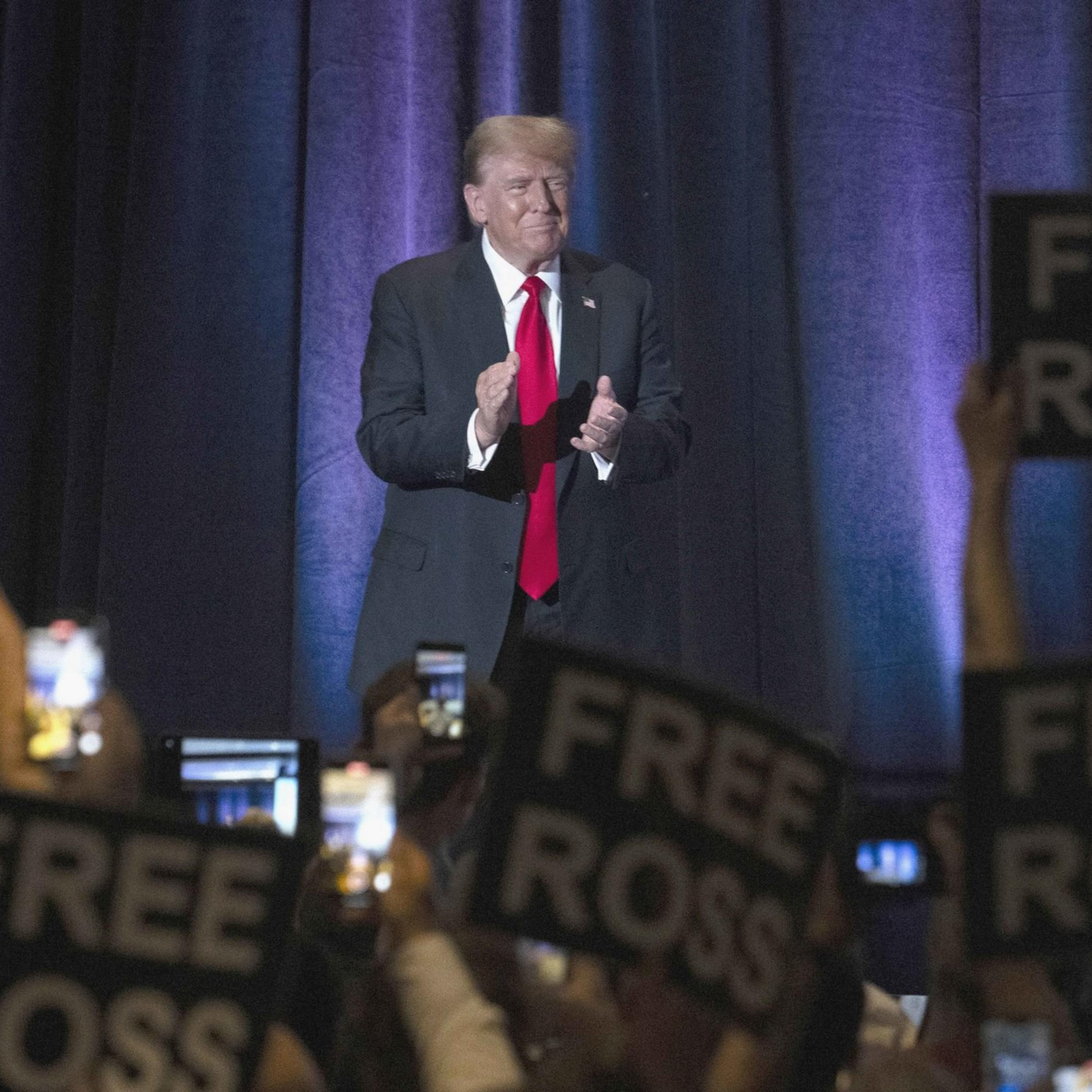 A man in a suit and red tie stands on a stage, clapping. The audience is holding up signs reading "FREE ROSS" and raising phones.