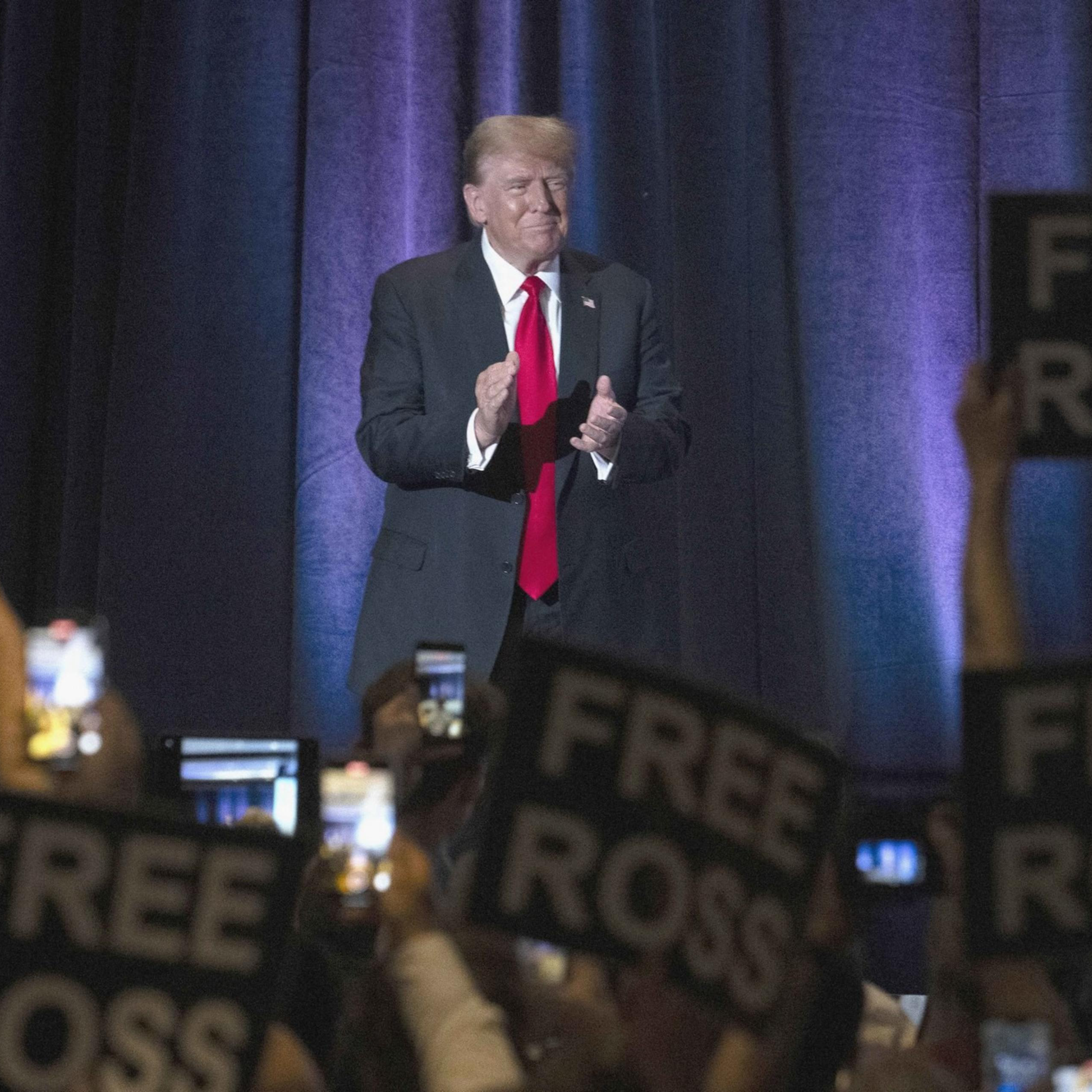 A man in a suit and red tie stands on a stage, clapping. The audience is holding up signs reading "FREE ROSS" and raising phones.