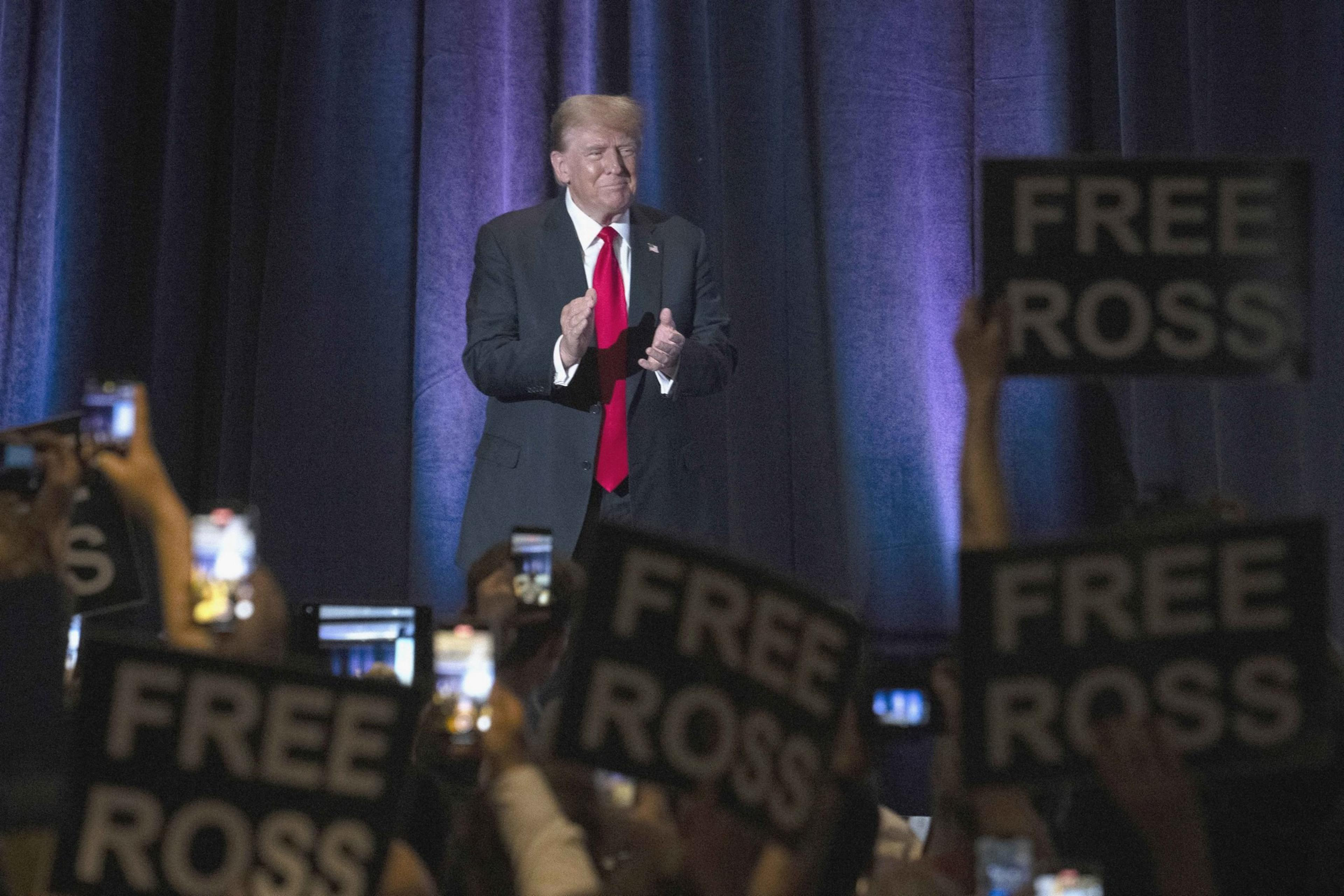A man in a suit and red tie stands on a stage, clapping. The audience is holding up signs reading "FREE ROSS" and raising phones.