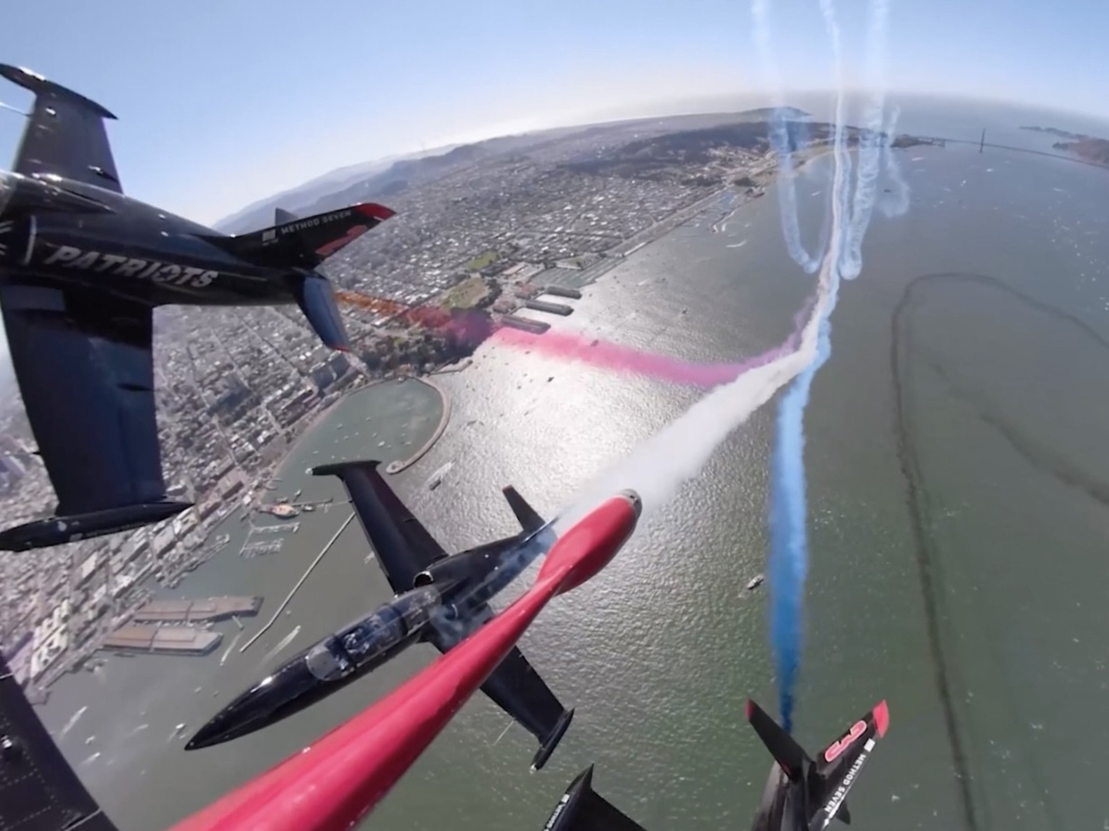 Four fighter jets fly in formation over a coastal city, releasing red, white, and blue smoke trails with water and bridges visible below.